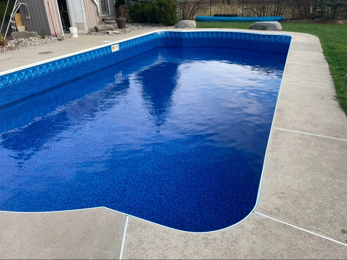 In-ground backyard swimming pool with blue interior, surrounded by concrete patio, with a grassy yard and trees in the background.