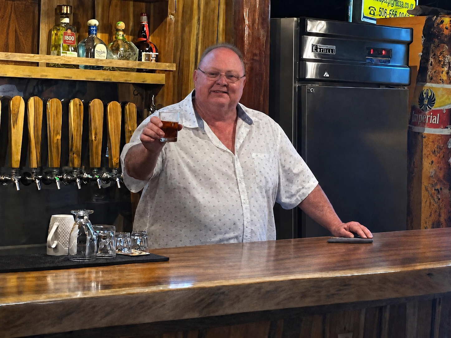 Sámara Brewing Company founder Tom Brinkerhoff holding a glass of house-brewed beer behind the wooden bar.