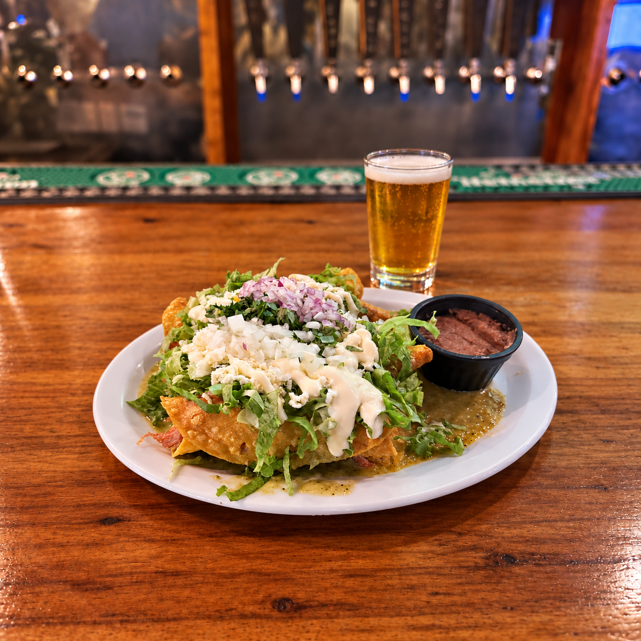 Plate of nachos topped with cheese, lettuce, onions, and sour cream, served with a side of beans, a glass of beer, and a dipping sauce, on a wooden bar counter with beer taps in the background.