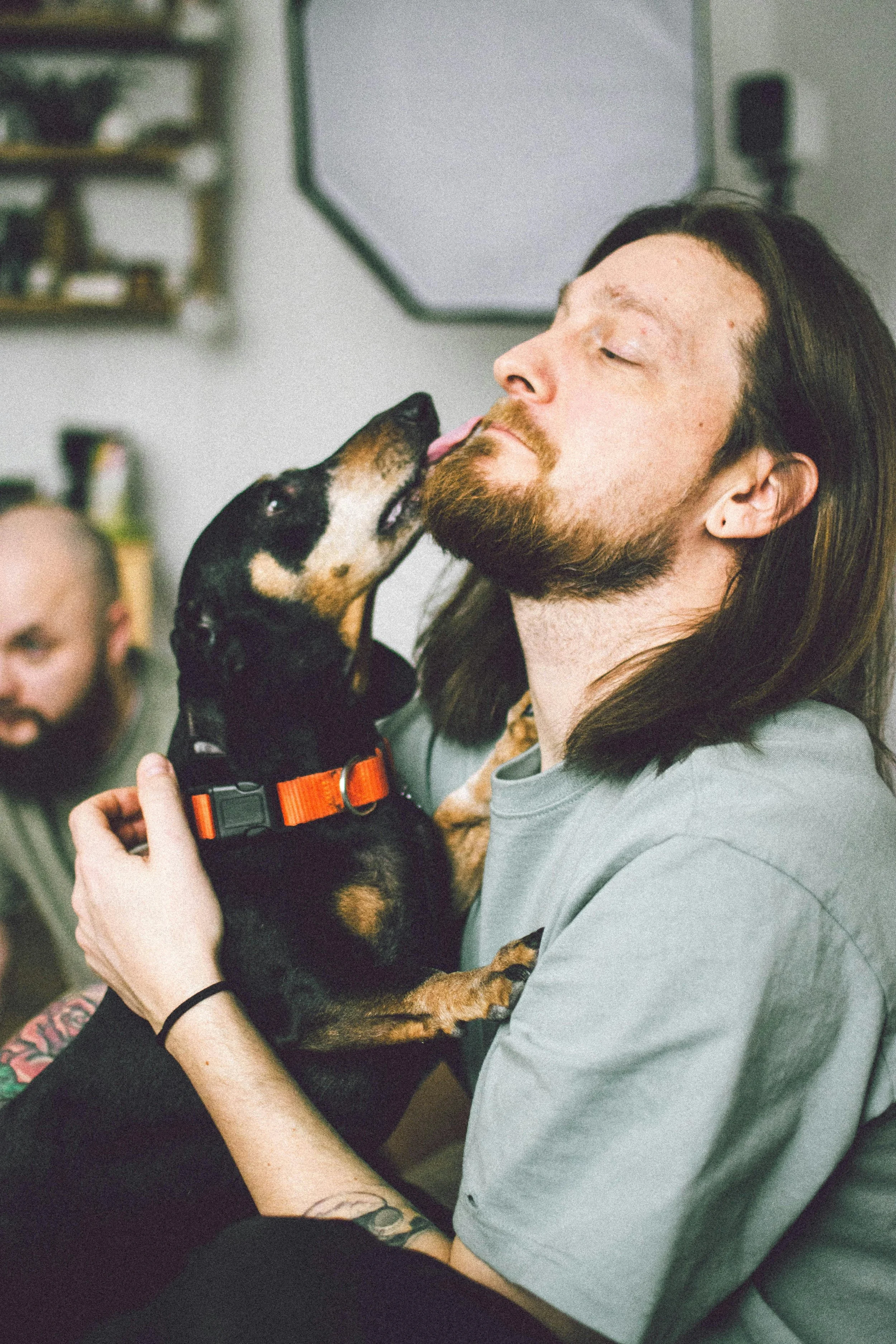 A man with long dark hair and a beard is holding a black and tan dog with a red collar, and the dog is licking his face affectionately.