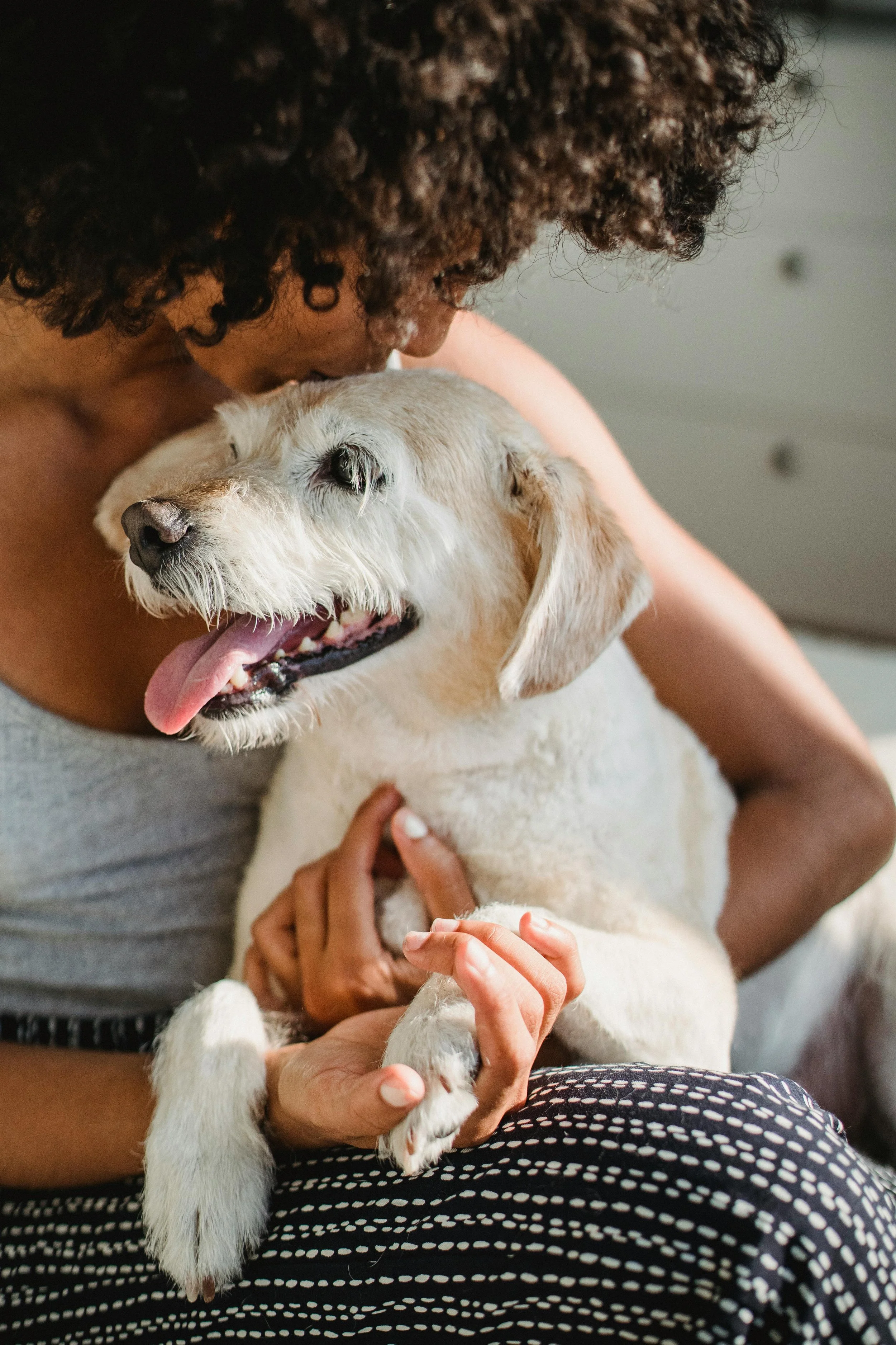 A woman with dark, curly hair is holding a small, light-colored dog with floppy ears and a pink tongue hanging out. The woman is close to the dog and appears to be nuzzling or kissing it. The dog looks happy and relaxed.