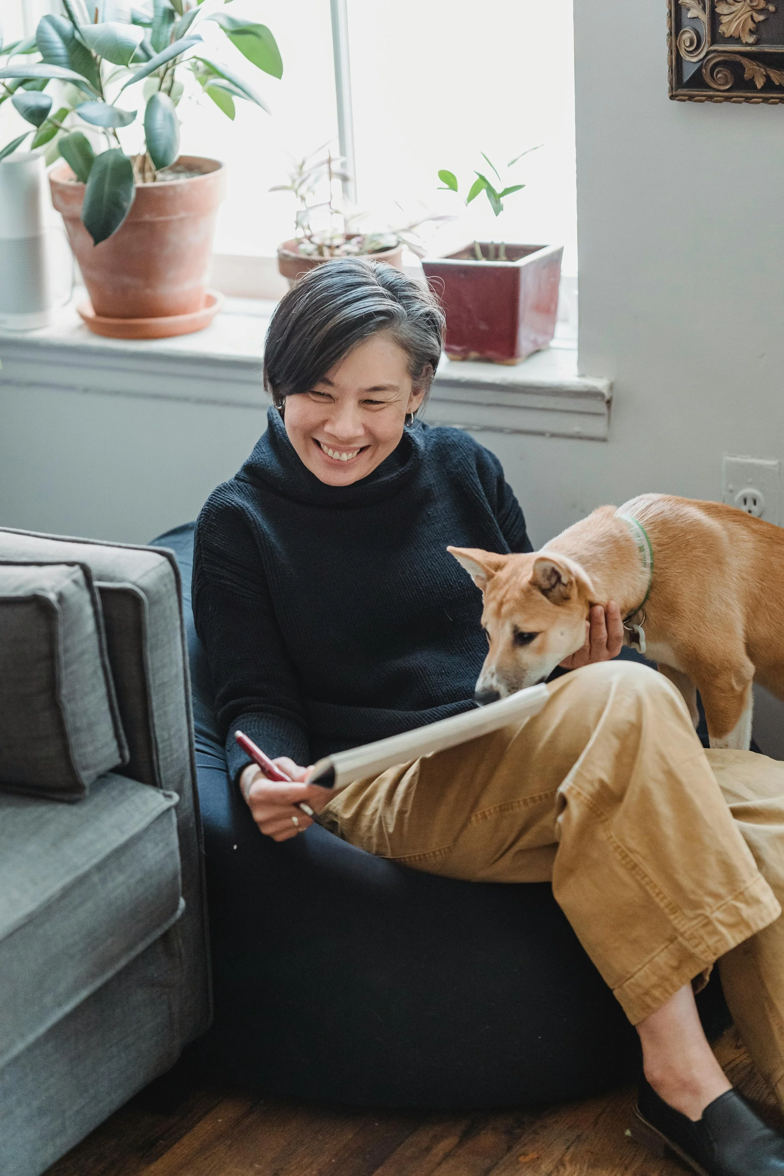 A woman with short dark hair smiling while sitting on the floor, holding a tablet, with a dog standing beside her. The woman is wearing a black sweater and tan pants, and the dog is tan with a green collar. In the background, there are potted plants on a windowsill and part of an ornate picture frame on the wall.