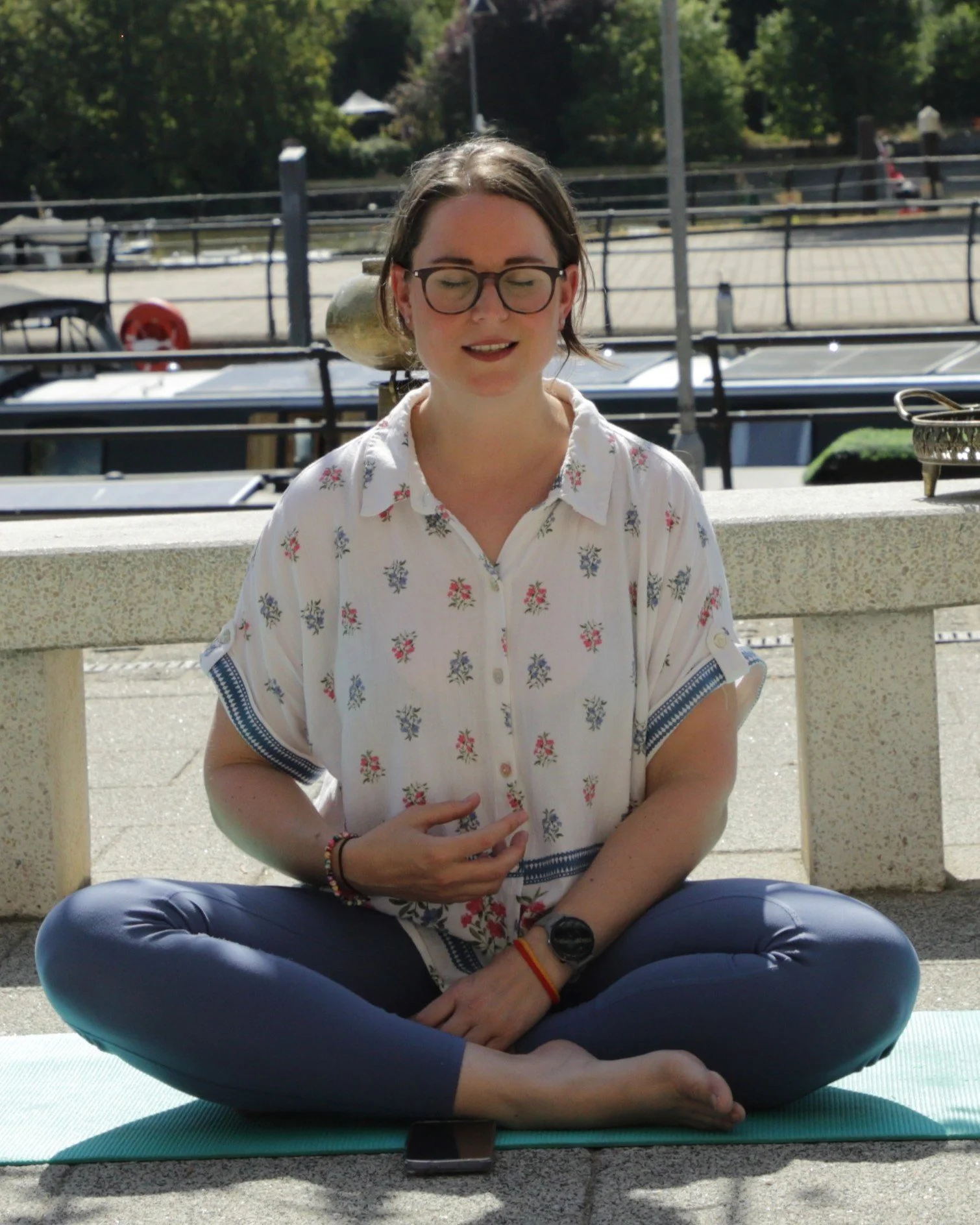 Jen, a white woman with brown tied-back hair and glasses, sits cross-legged outside in blue leggings and a white loose shirt with blue accents. She has her eyes closed and gestures gently towards her middle in a guided mindfulness practice.