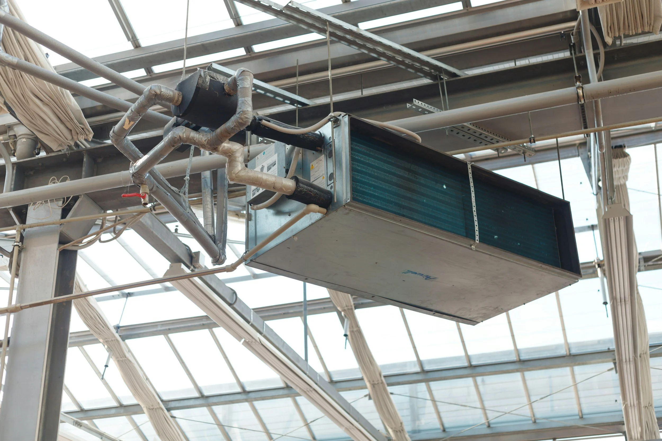 Large HVAC air conditioning unit mounted under a glass ceiling with visible pipes and wiring.