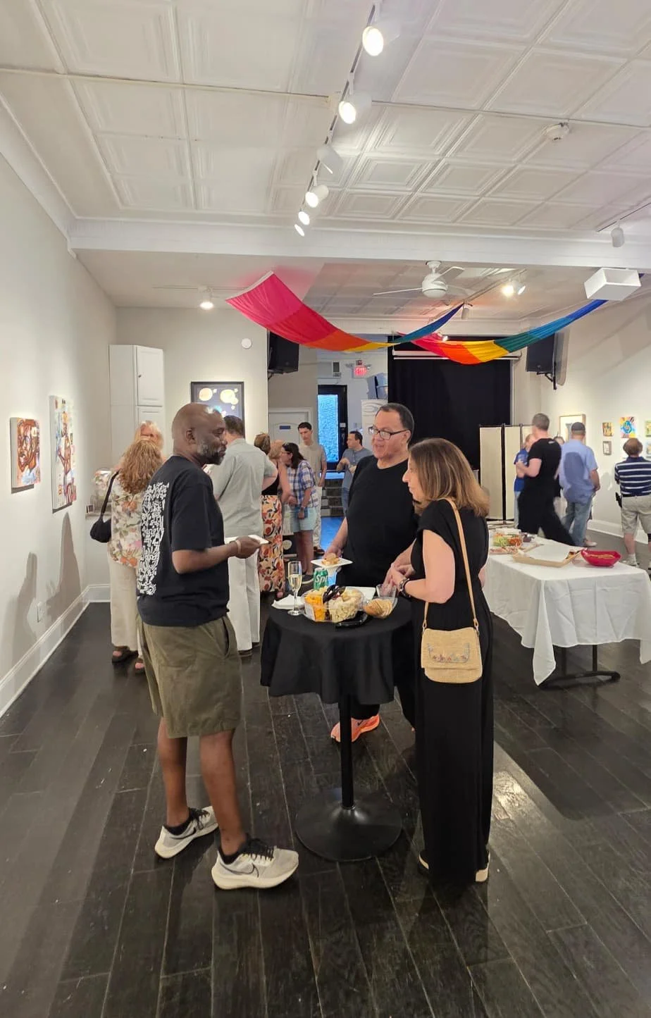 People mingling and eating at a social event inside an art gallery with colorful decorations hanging from the ceiling.