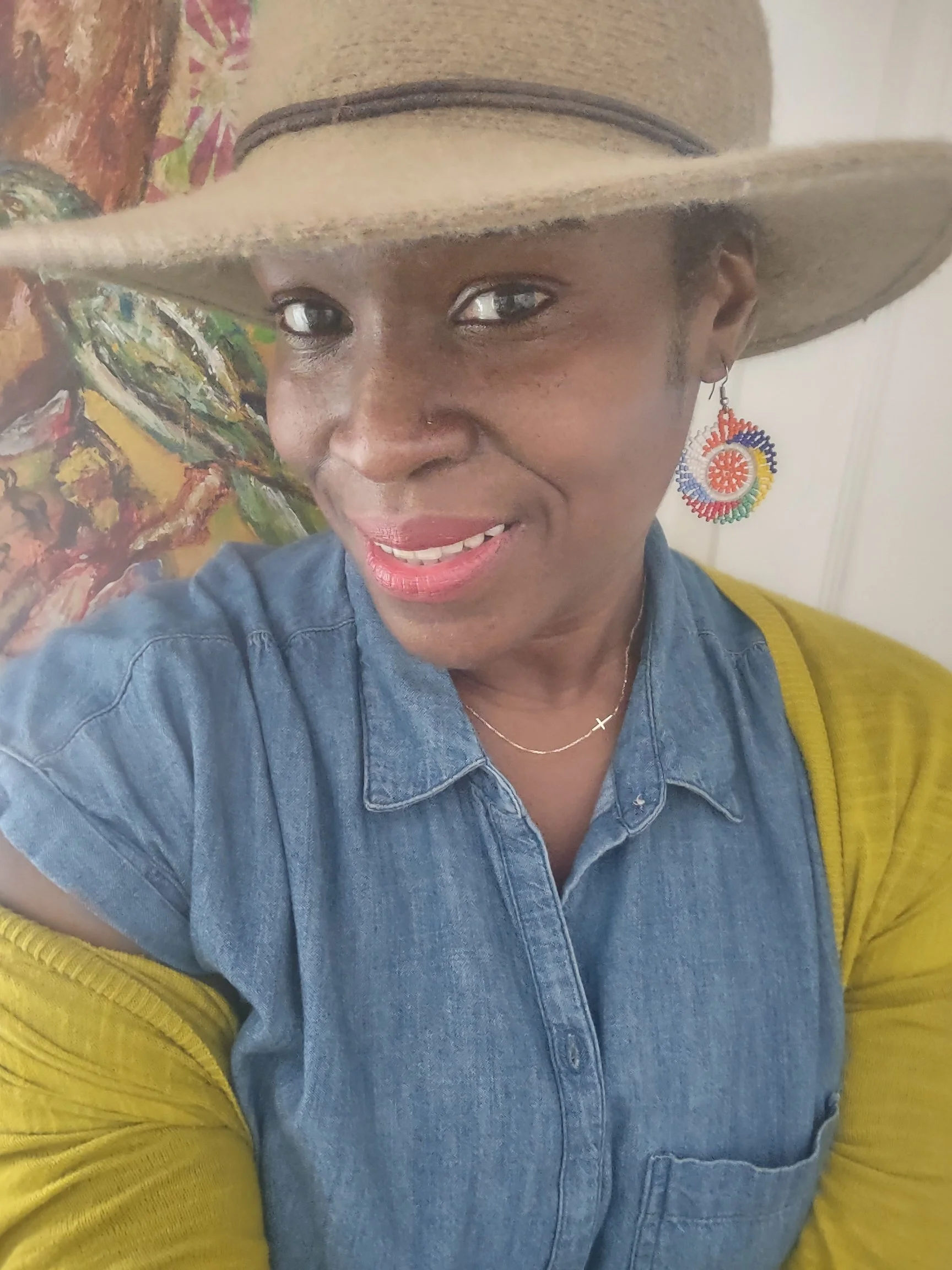 A woman wearing a large tan cowboy hat, colorful spiral earring, denim shirt, yellow cardigan, and a small cross necklace, smiling at the camera with an abstract colorful painting in the background.
