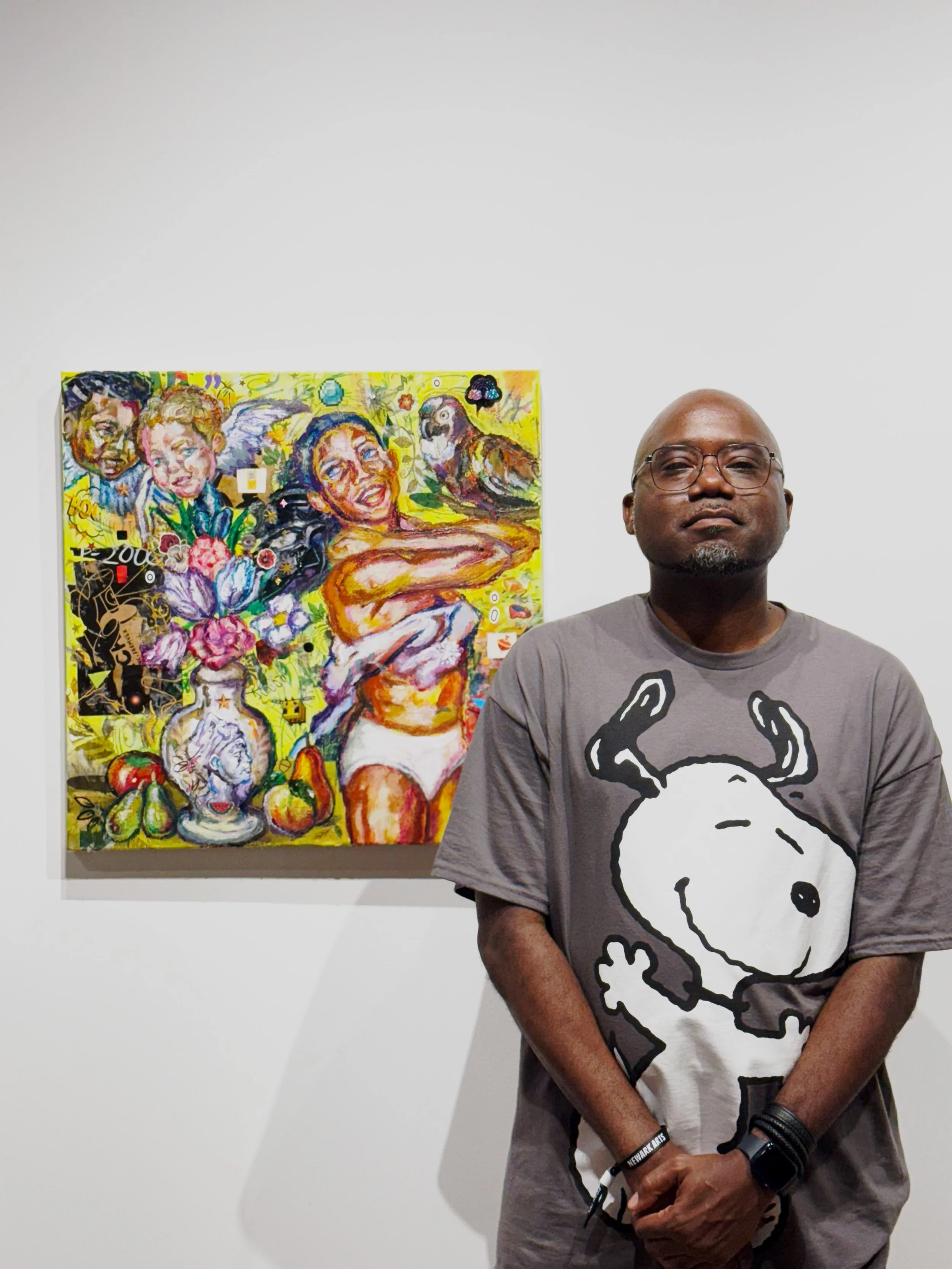 A man with glasses and a gray t-shirt featuring Snoopy, standing in front of a colorful, graffiti-style painting of multiple faces and various abstract elements, on a white gallery wall.