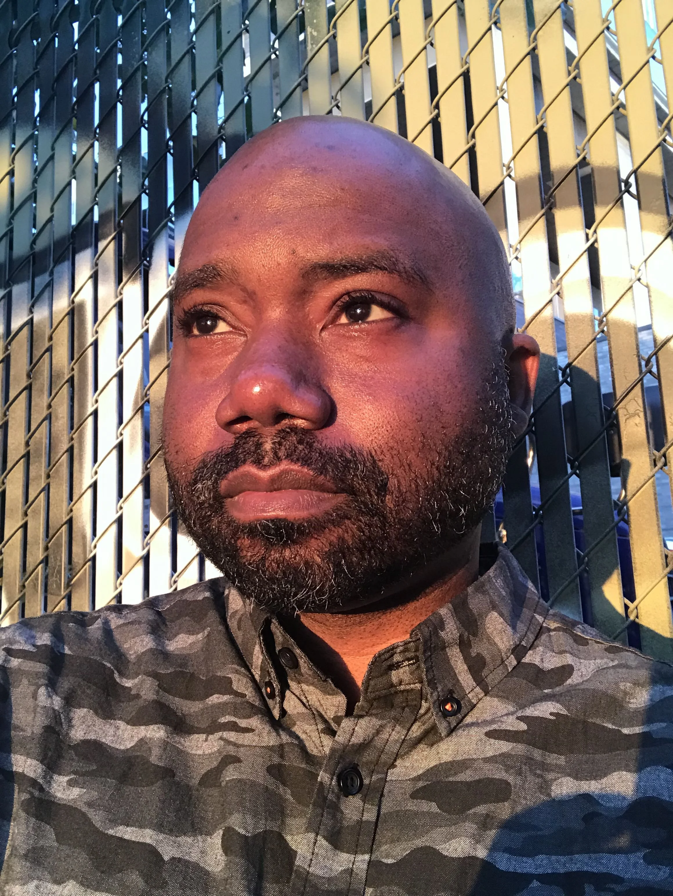 Close-up of a man with a shaved head and beard, standing against a chain-link fence.