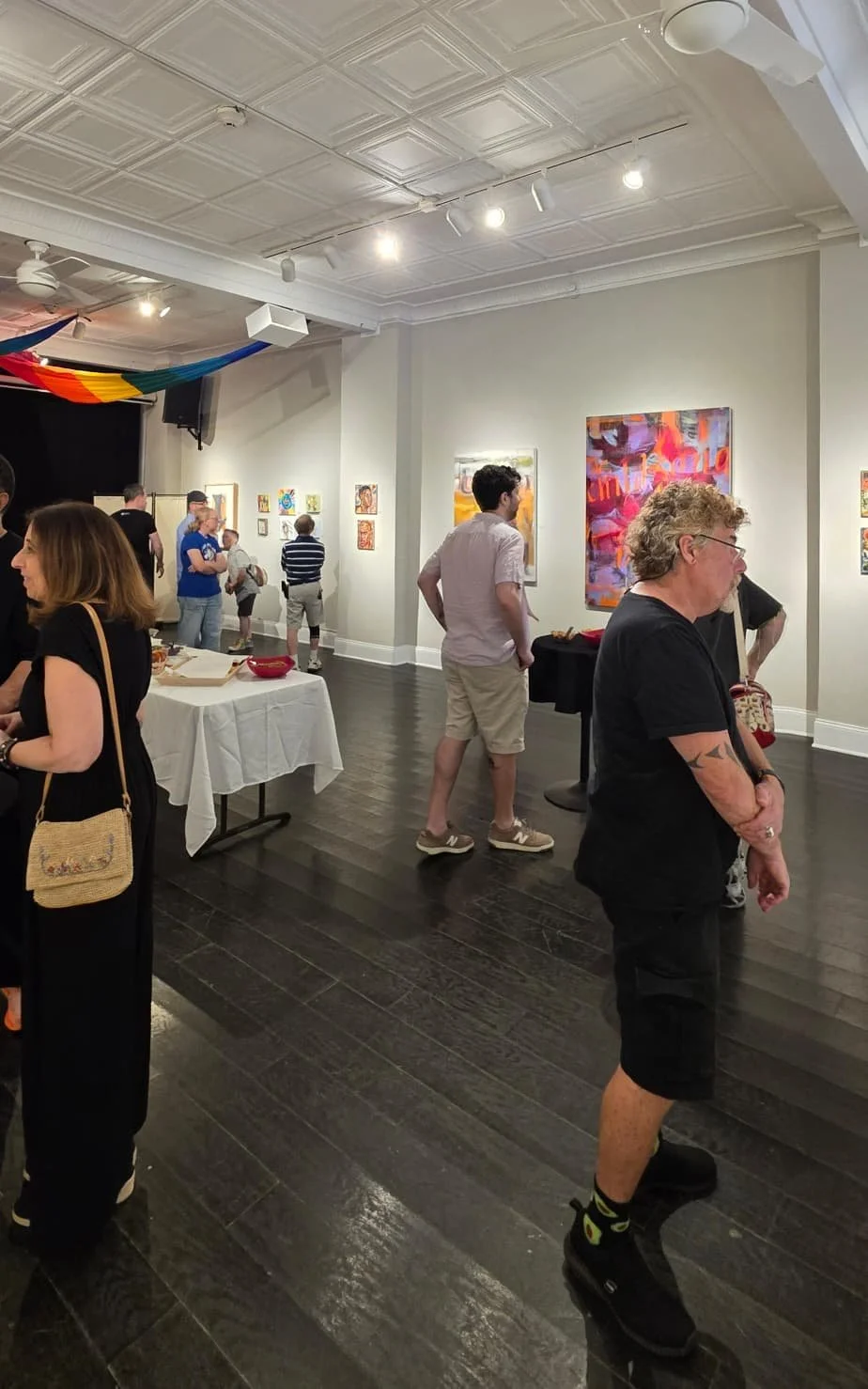 People viewing colorful abstract paintings in an art gallery with white walls, dark wood floors, and ceiling lights.