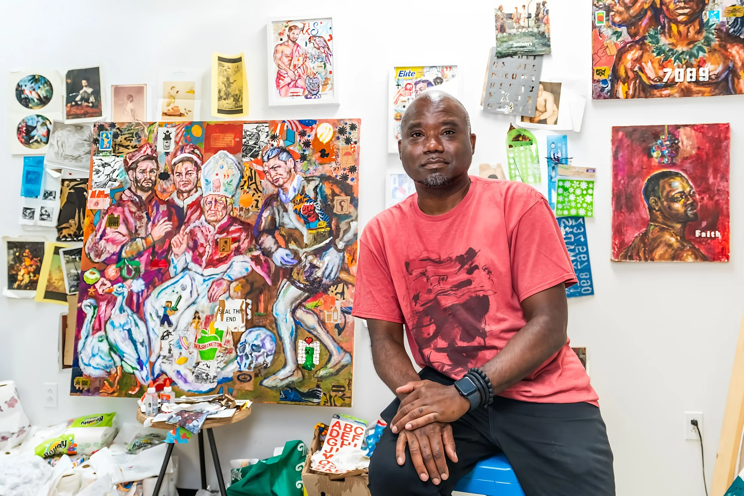 An artist wearing a red t-shirt and a black smartwatch is sitting on a blue stool in his art studio. The studio has multiple colorful paintings on the white wall behind him, including portraits and abstract art. Art supplies and papers are scattered on a small table in front of him.