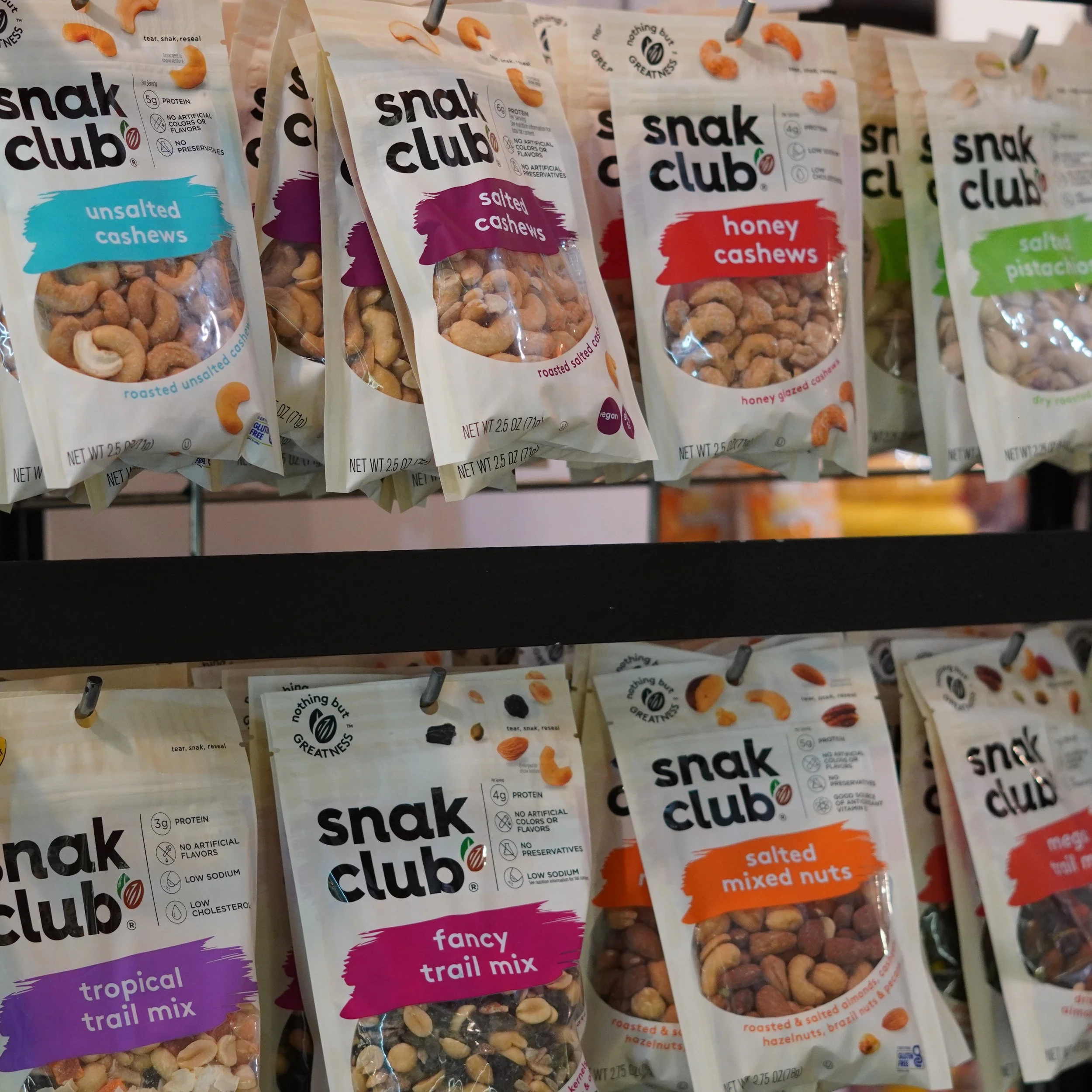 Shelves of various Snak Club roasted and flavored nuts including unsalted cashews, salted cashews, honey cashews, fancy trail mix, salted mixed nuts, and tropical trail mix in snack-sized bags at The French Kitchen 4th floor, Westin Bonaventure