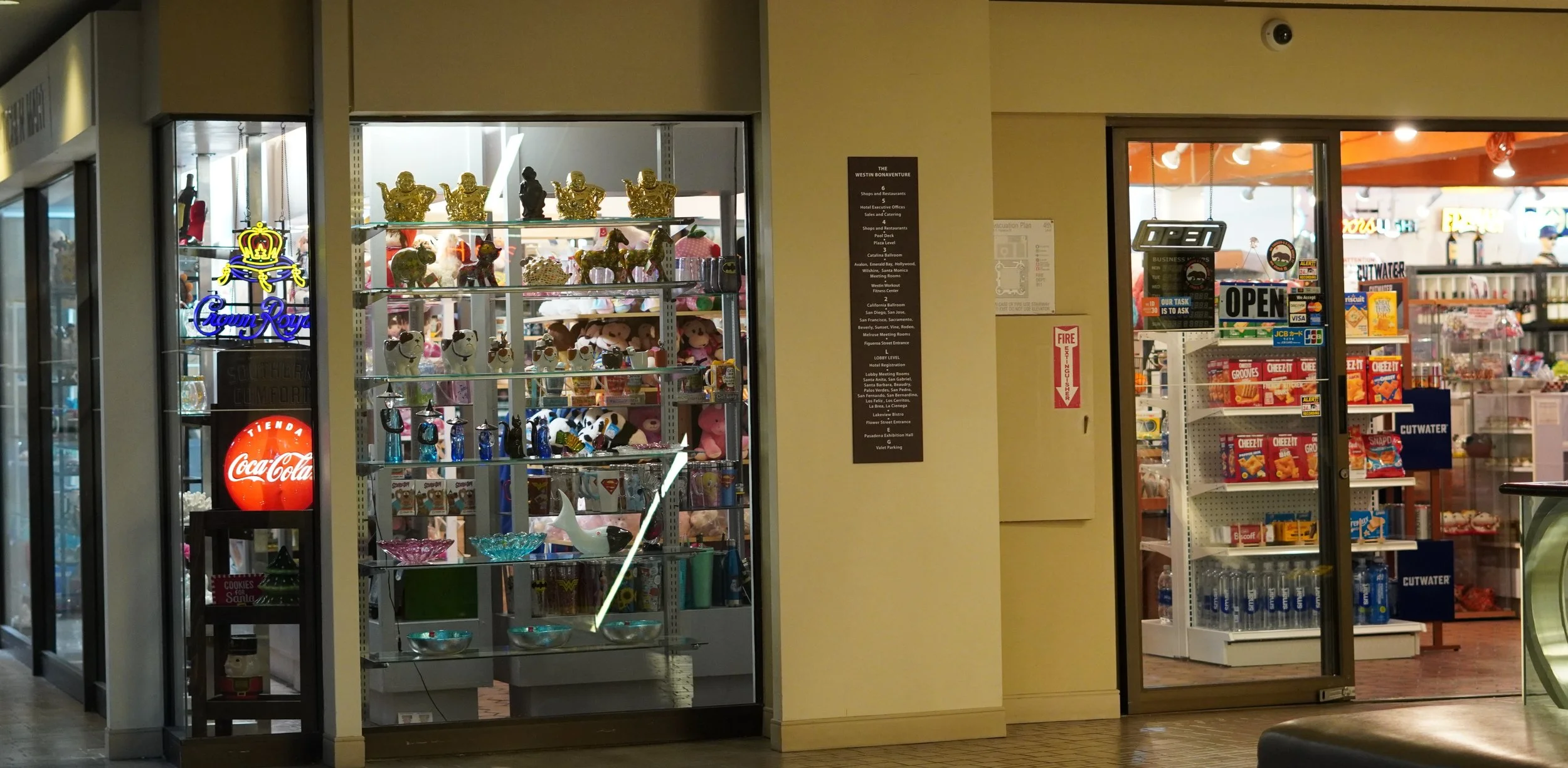 Inside a shopping mall, a storefront displays various decorative items like figurines, plush toys, and glassware, with a glass door entrance to a snack shop selling chips and bottled drinks nearby.