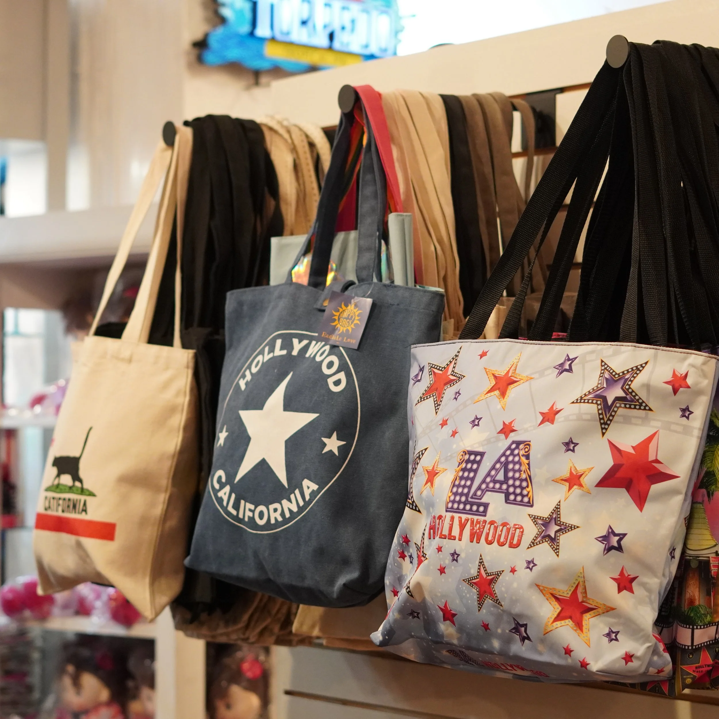 Various tote bags hanging on display, including a beige tote with a black cat graphic and the word California, a dark gray tote with a white Hollywood sign and star design, and a colorful tote with stars and the text 'LA Hollywood'. available at The 