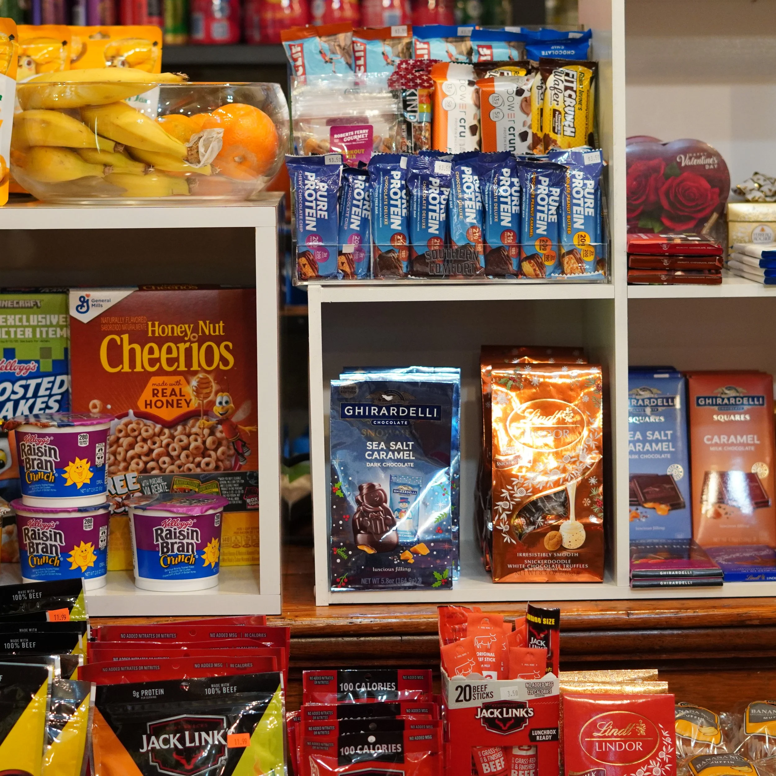 Grocery store shelf displaying various snacks, candies, and cereals, including Cheerio Honey Nut cereal, Ghirardelli chocolate, Lindt Lindor truffles, Jack Link's beef jerky, and fruit in a glass bowl at The French Kitchen 4th floor Westin Bonaventur