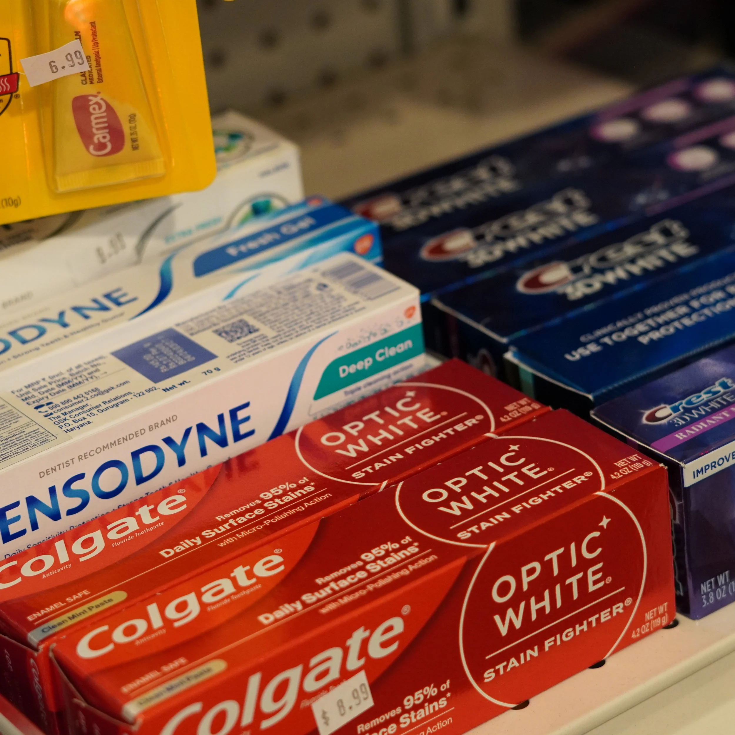 Shelf display of oral care products including Colgate Optic White stain fighter toothpaste, Sensodyne Deep Clean toothpaste, and Crest whitening strips.