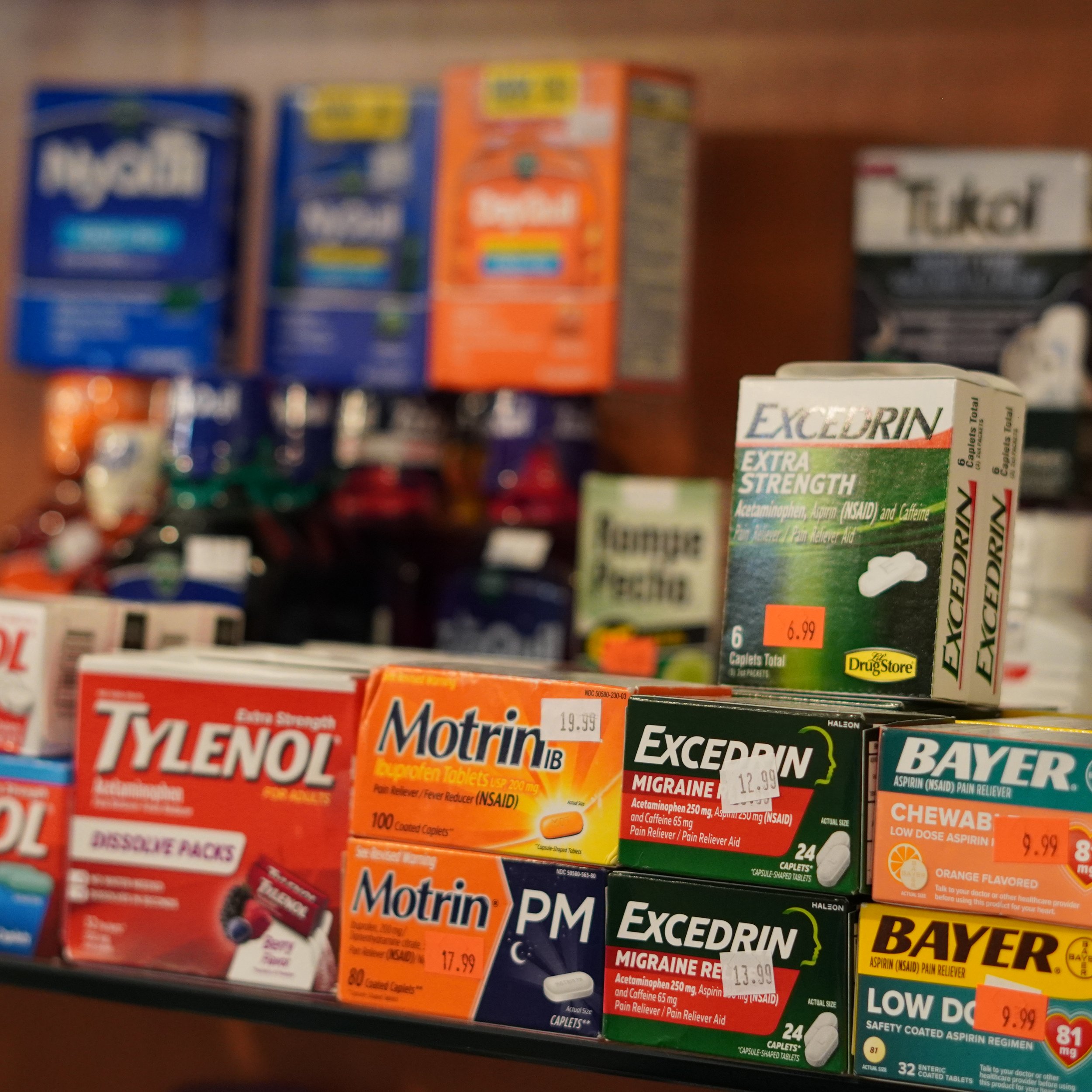 Over-the-counter medication boxes on store shelves, including Tylenol, Motrin, Excedrin, Bayer, and Taked, with price tags visible.