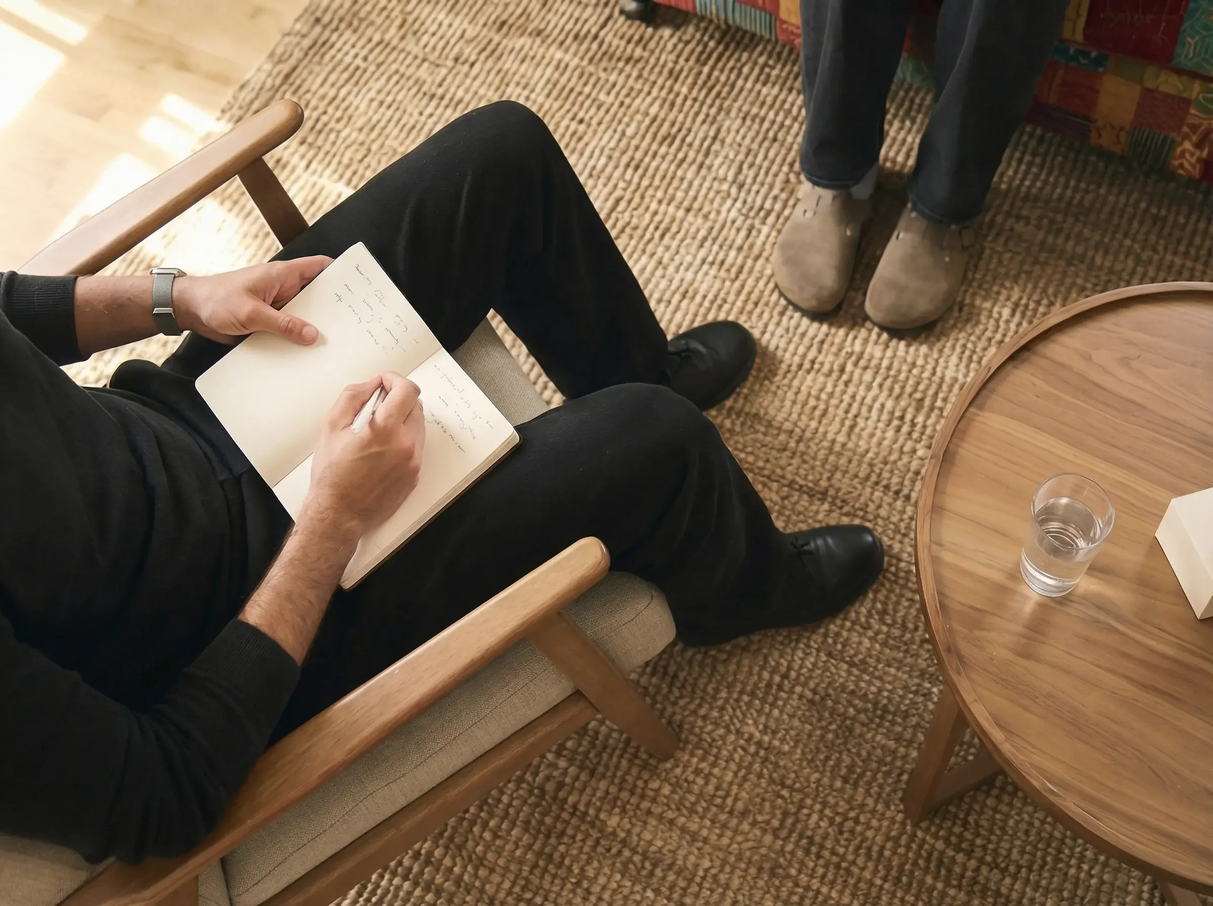 Person sitzt auf einem Stuhl, schreibt in einem Notizbuch, während eine andere Person mit braunen Schuhen steht, neben einem runden Holztisch mit einem Glas Wasser.