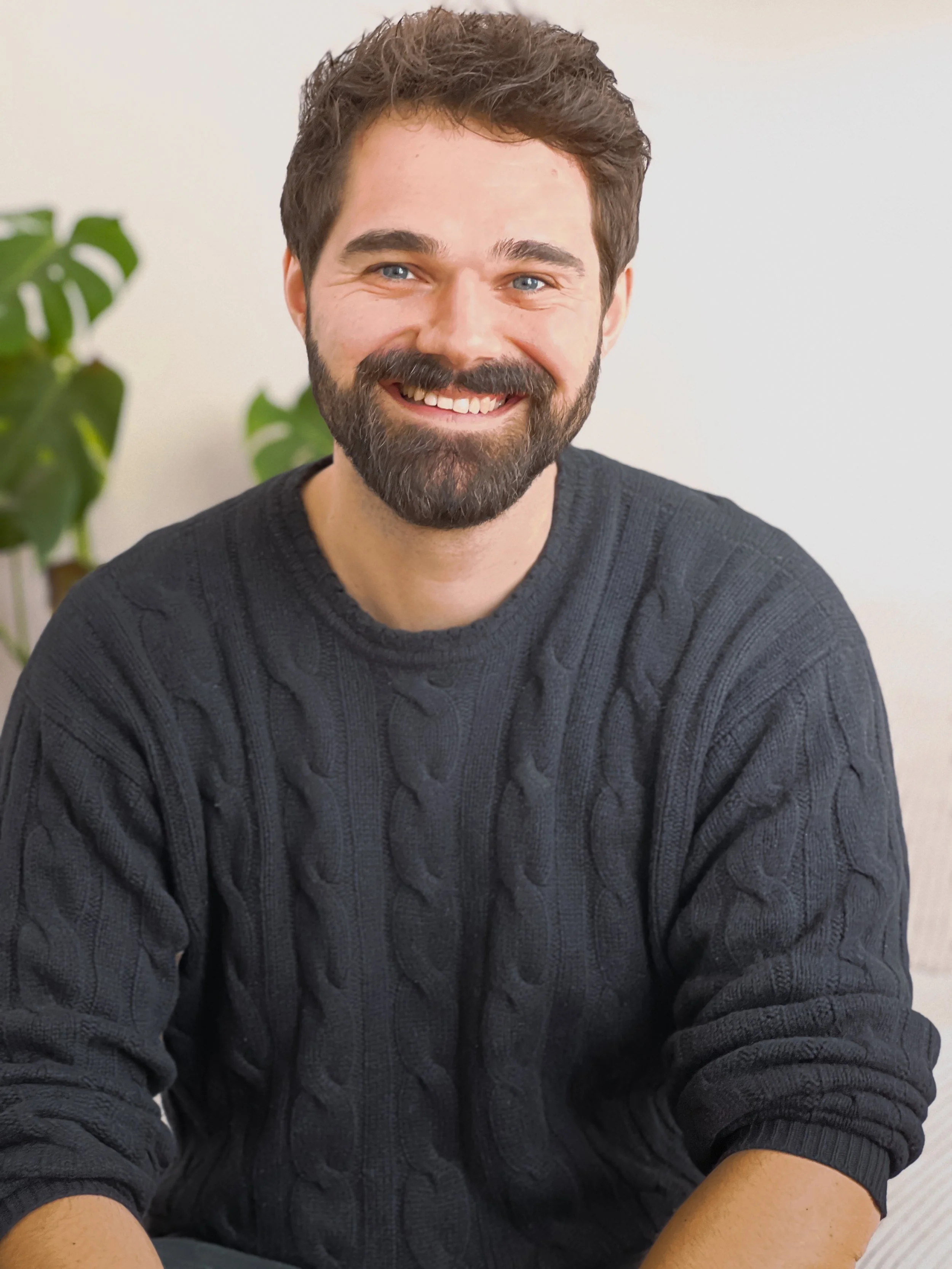Ein lachender Mann mit dunklem Haar und Bart, trägt einen schwarzen Wollpullover, sitzt in einem Raum mit weißen Wänden und einer Pflanze im Hintergrund.