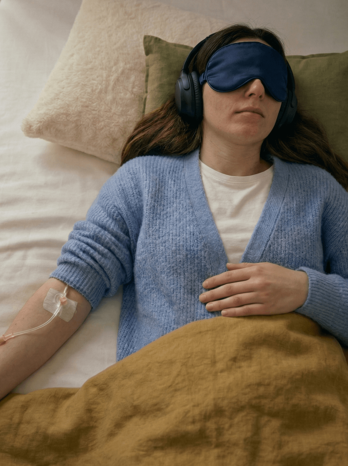 Eine Frau liegt im Bett, trägt eine Schlafmaske und hört Musik mit Kopfhörern. Sie hat eine Infusionsnadel in den Arm.