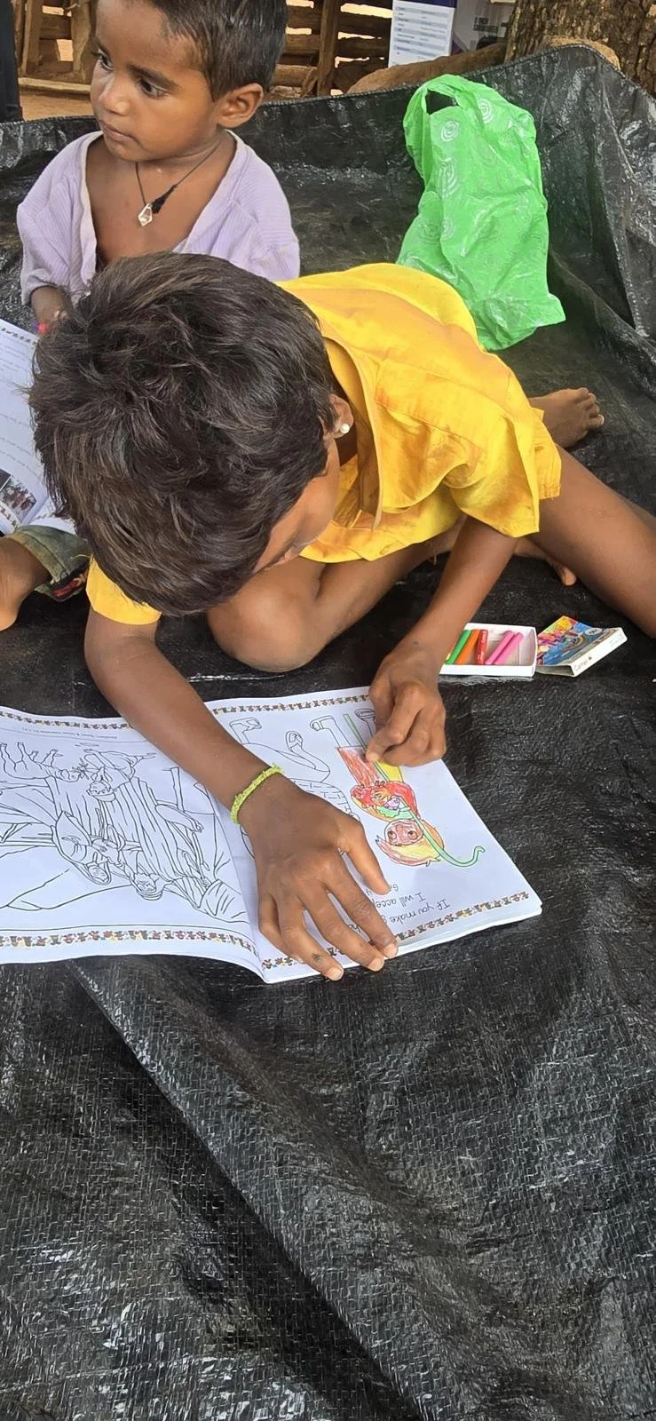 Two children sitting on black plastic sheet, one coloring a picture of a princess with crayons, the other observing, with a green plastic bag in the background.