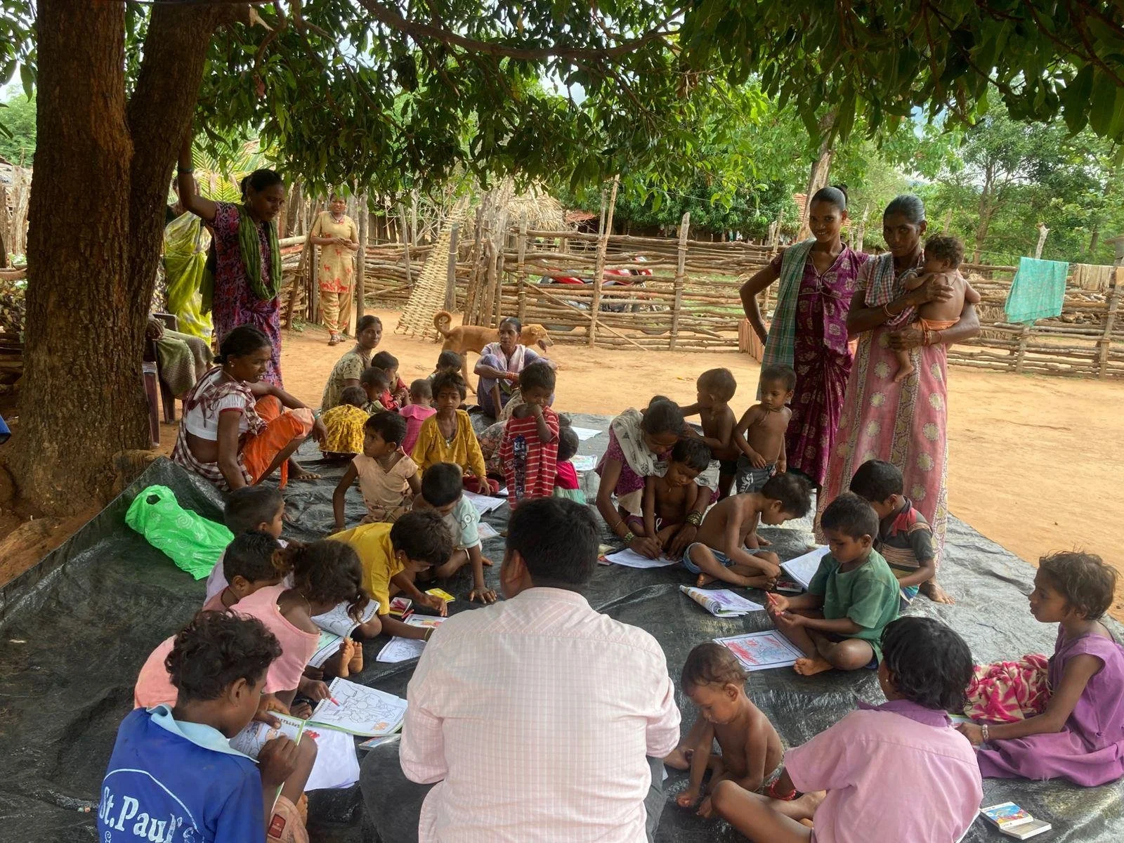Children and adults gathered outdoors under a large tree, engaged in drawing and reading activities on a black tarp, with a rustic wooden fence and trees in the background.