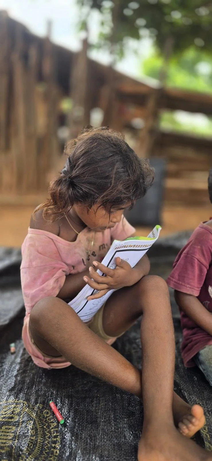 A young girl with curly hair sitting cross-legged, looking at a workbook, in an outdoor setting with a wooden fence and greenery in the background.