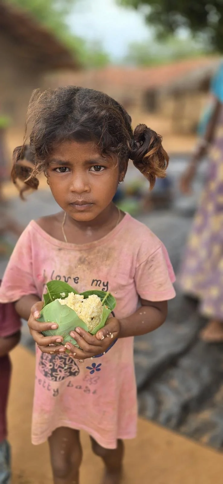 A young girl with messy hair and a serious expression, holding a leaf with a food item inside, in an outdoor setting.