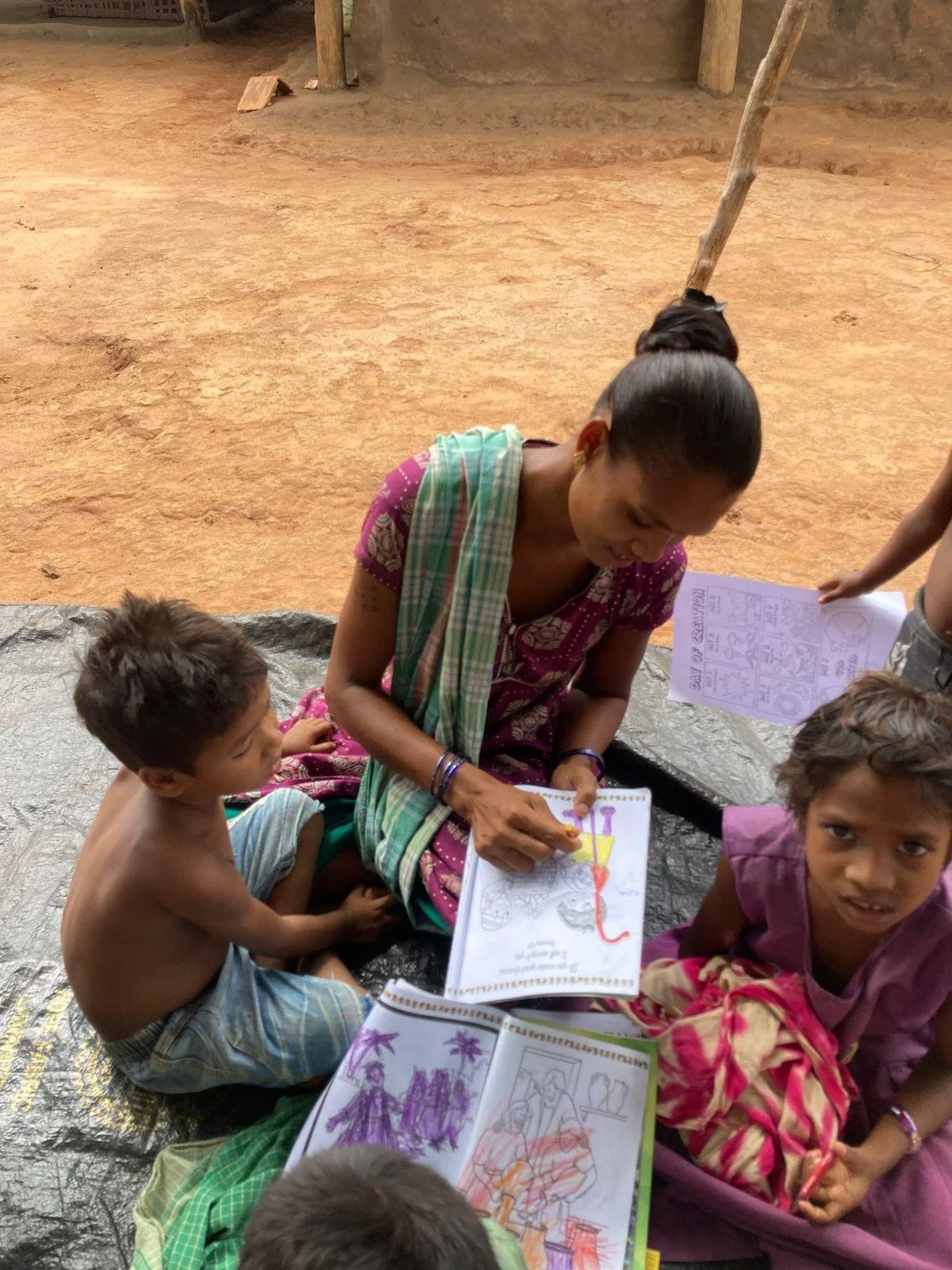 Woman and children sitting on ground reading and looking at coloring books outside.