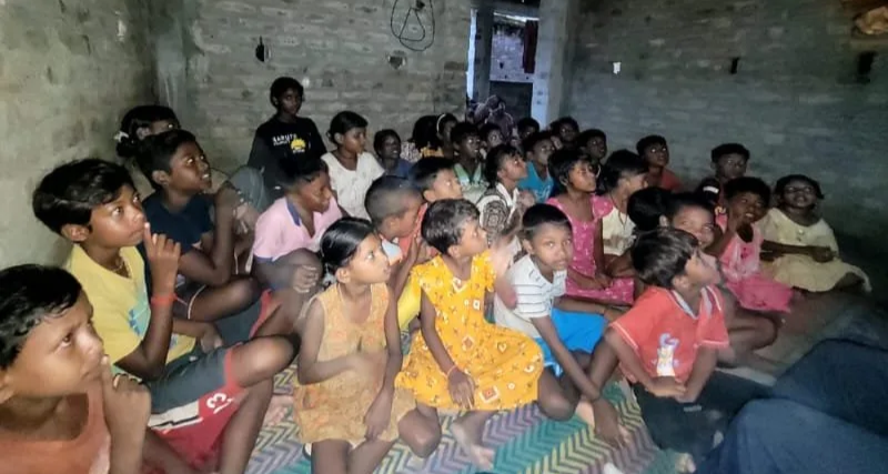 Group of children sitting on the floor inside a rustic, unfinished brick room, attentively watching something out of the frame.