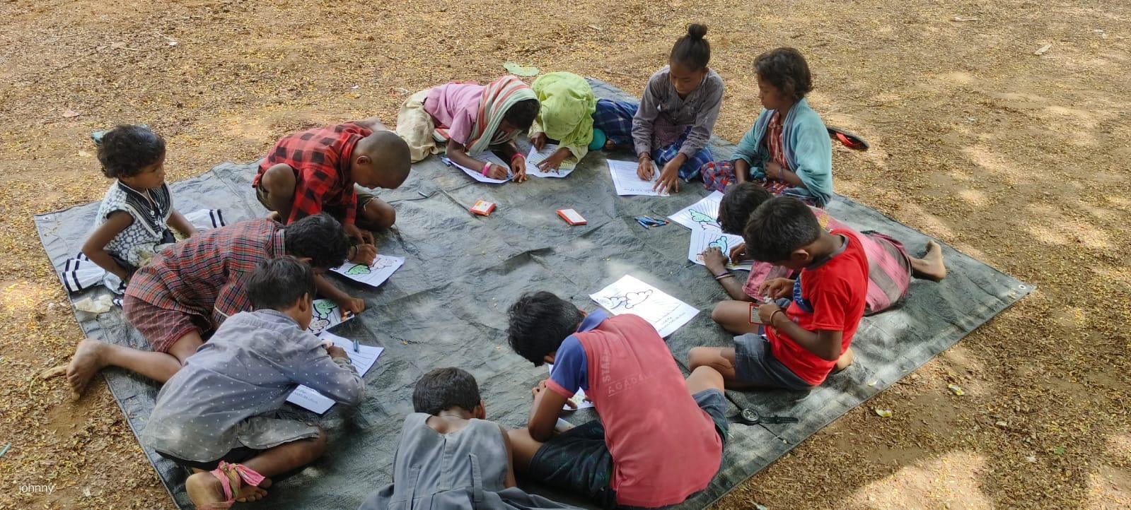 Children and a woman are sitting on a tarp on the ground outdoors, engaged in drawing or coloring activities with papers and coloring materials.