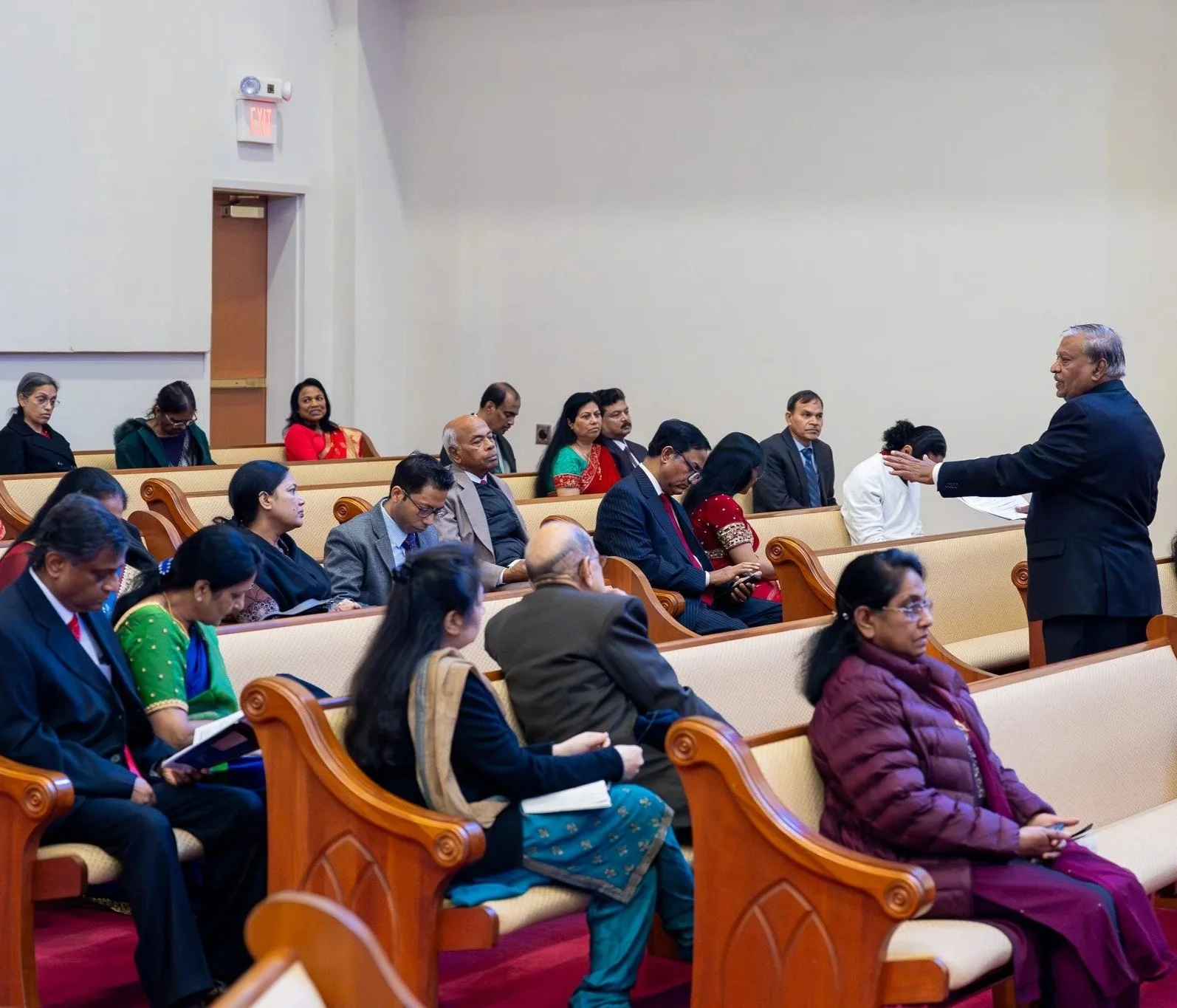 A man in a dark suit is speaking to an audience in a church or conference hall. The audience, consisting of men and women of diverse backgrounds, are seated on wooden benches with beige cushions, listening attentively. Some audience members are taking notes or looking at a book.