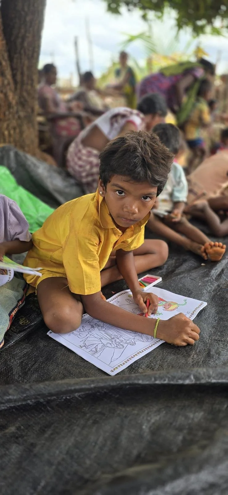 Young boy in yellow shirt sitting on ground, coloring a picture of a snake with crayons, outside with other children and adults in the background.