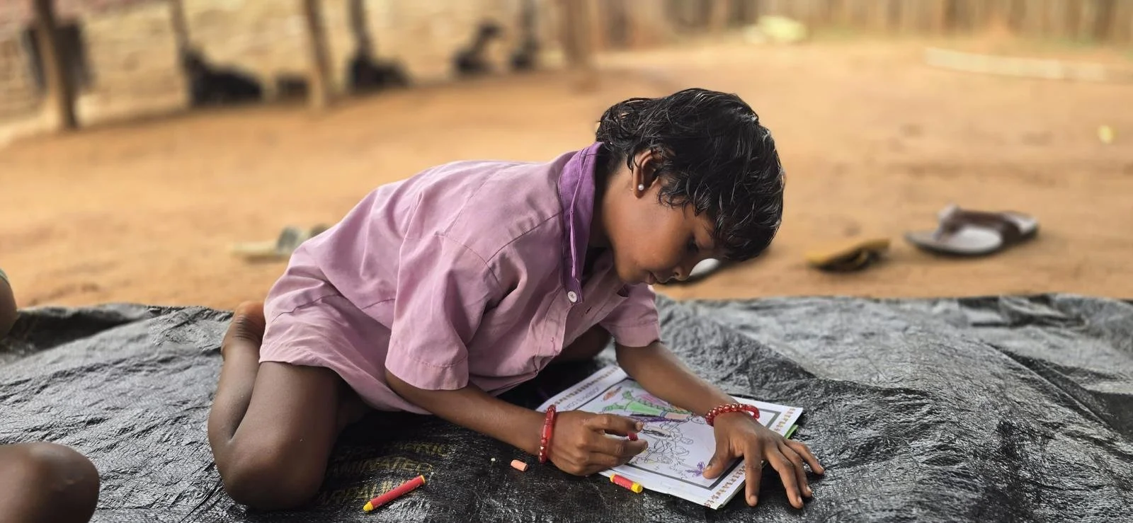 A young boy wearing a pink shirt, sitting on a black mat outdoors, engaged in coloring in a book with crayons, with a blurred background of a dirt ground and some shoes.