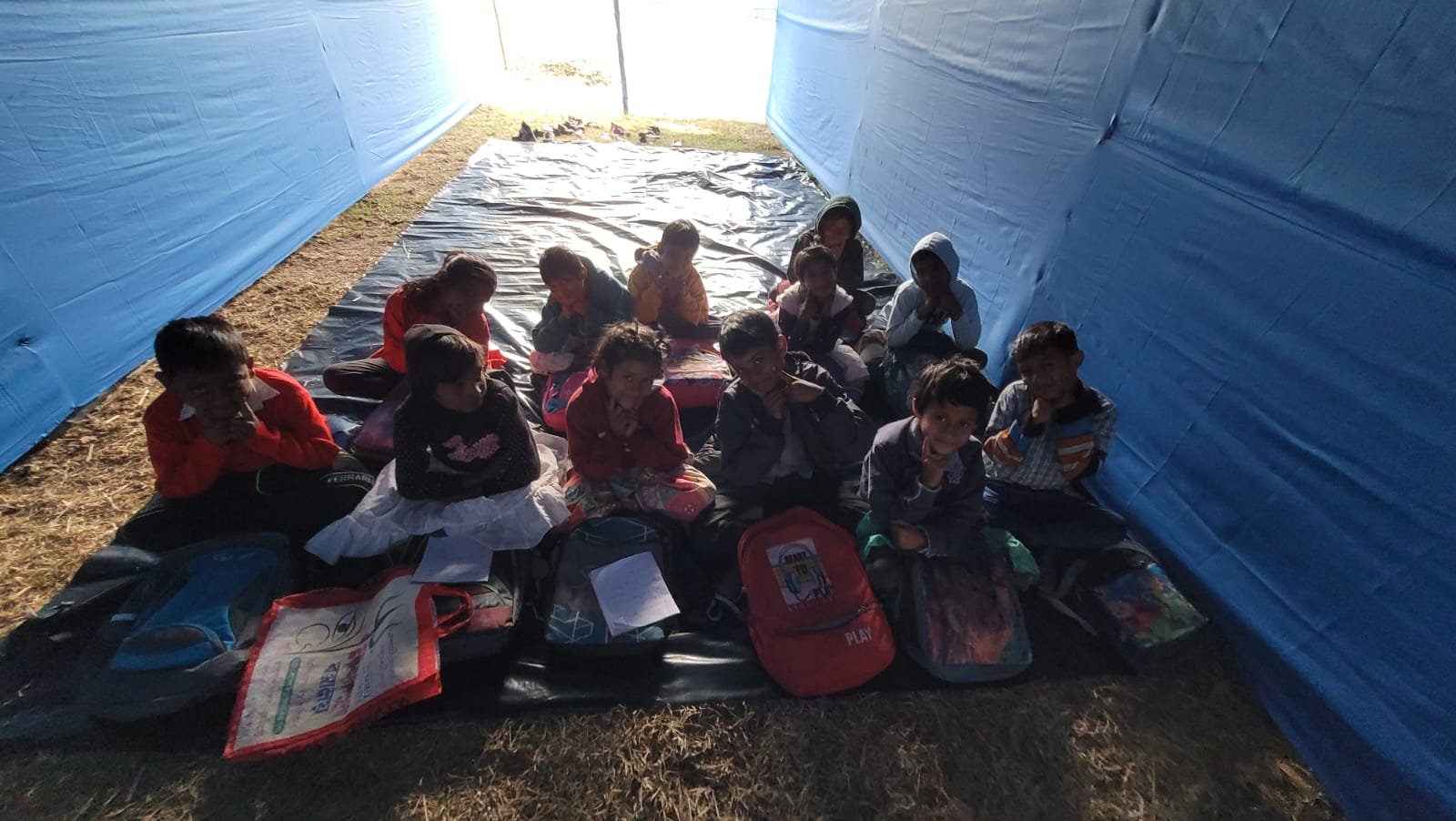 A group of children sitting on the ground inside a makeshift tent with blue walls, some with backpacks and books, during daytime.