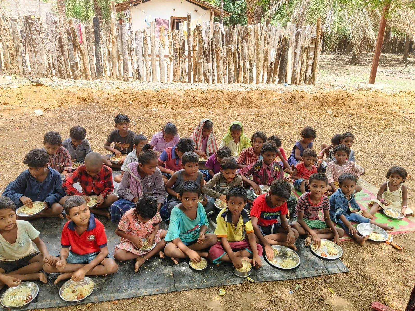 Group of children sitting outdoors on mats, eating a meal from plates, with a simple rural background featuring a wooden fence and trees.