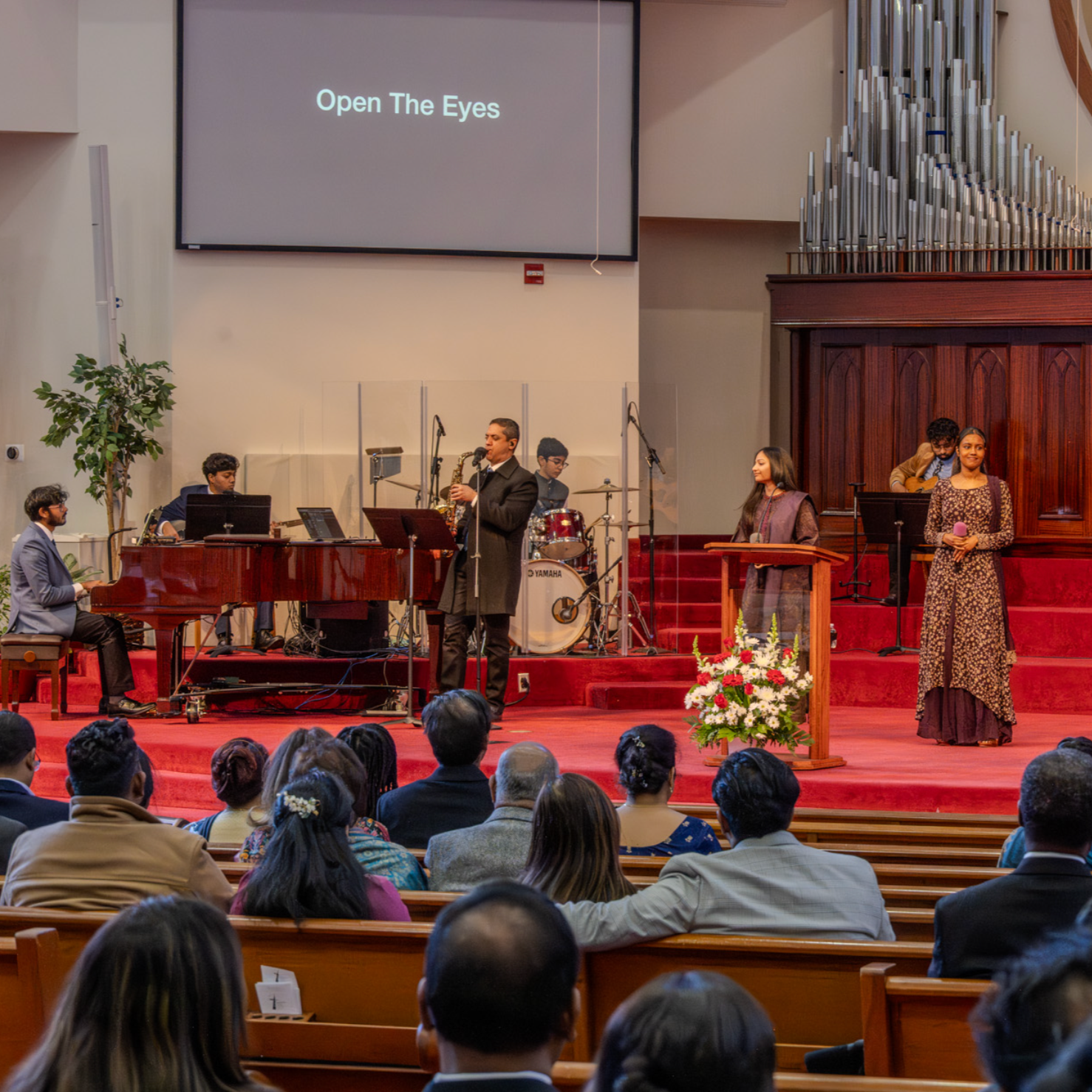 A musical performance taking place in a church or auditorium with a band and singers on a red carpeted stage. The band includes a pianist, saxophonist, drummer, and keyboardist. Two women are standing at the front, one singing into a microphone, and another playing a tambourine. The audience is seated facing the stage, and a large floral arrangement decorates the podium. A screen in the background displays the words "Open The Eyes."