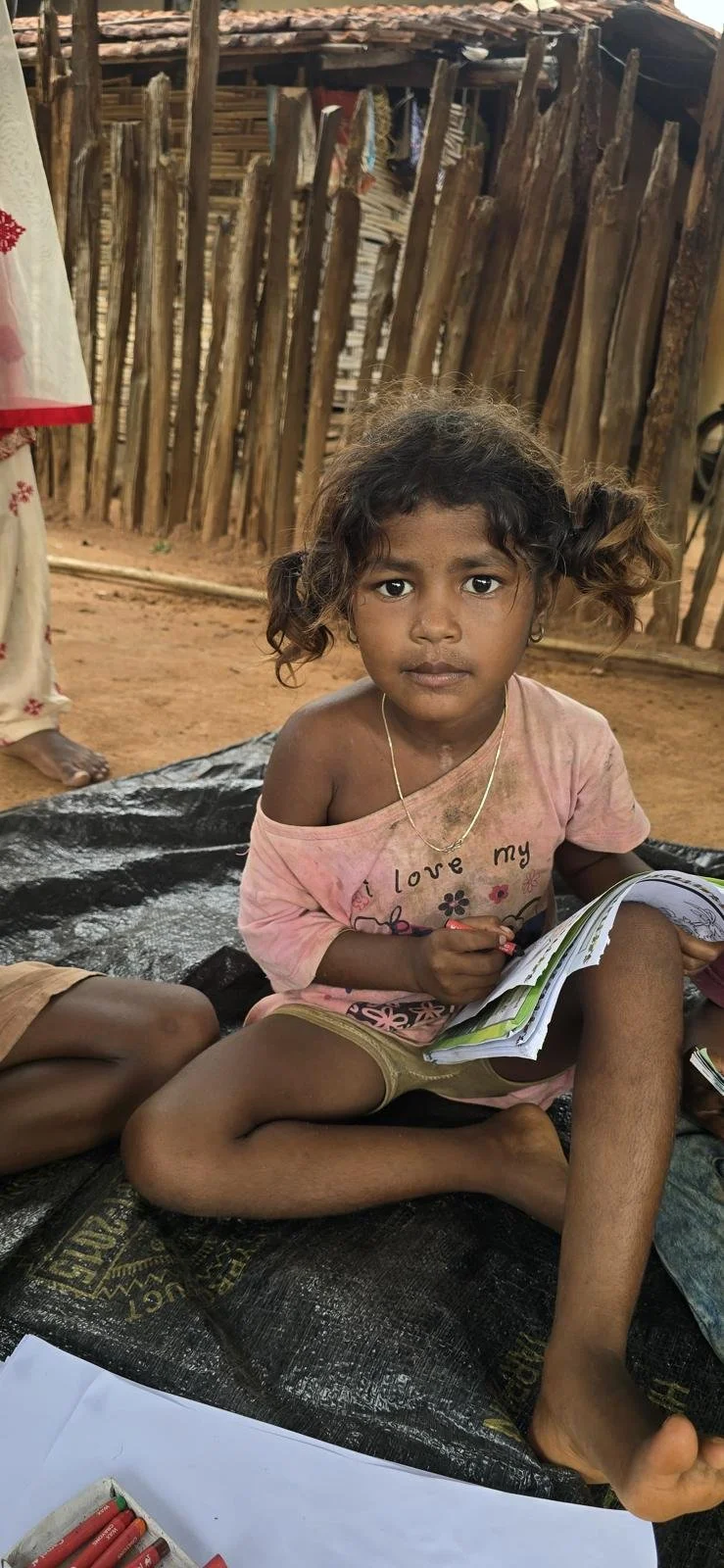 A young girl with curly hair sitting on a black plastic sheet with a book in her lap, looking directly at the camera. She is wearing a pink shirt with flowers and shorts, with a dirt smudge on her face and arm. The background shows a wooden fence and