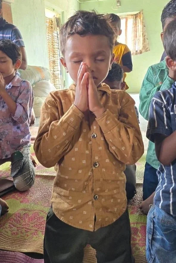 Young boy with short hair, wearing a brown shirt, praying with eyes closed and hands pressed together in a prayer pose, other children in the background also praying in a room with green walls and windows.