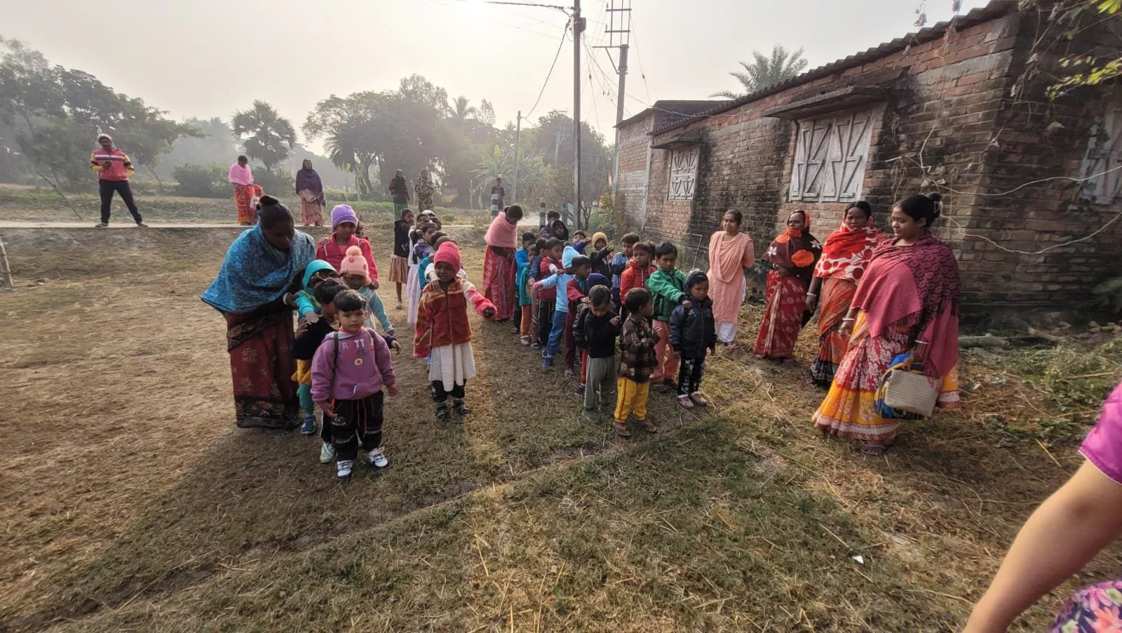 Children and adults standing in line outdoors, wearing jackets and warm clothing, near a brick building and trees in the background.