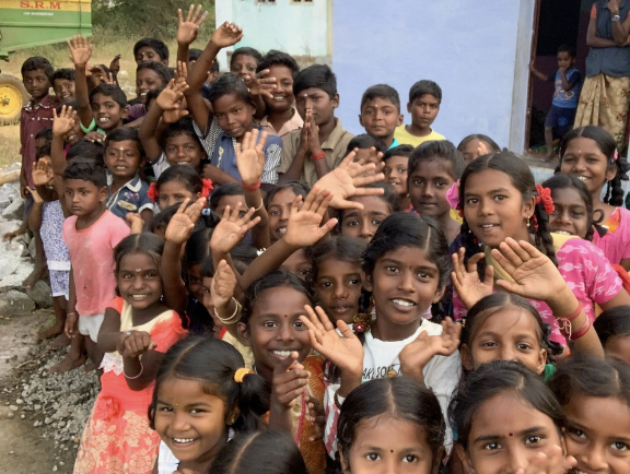 A large group of smiling children of diverse ages and skin tones, waving and looking at the camera outside a building.