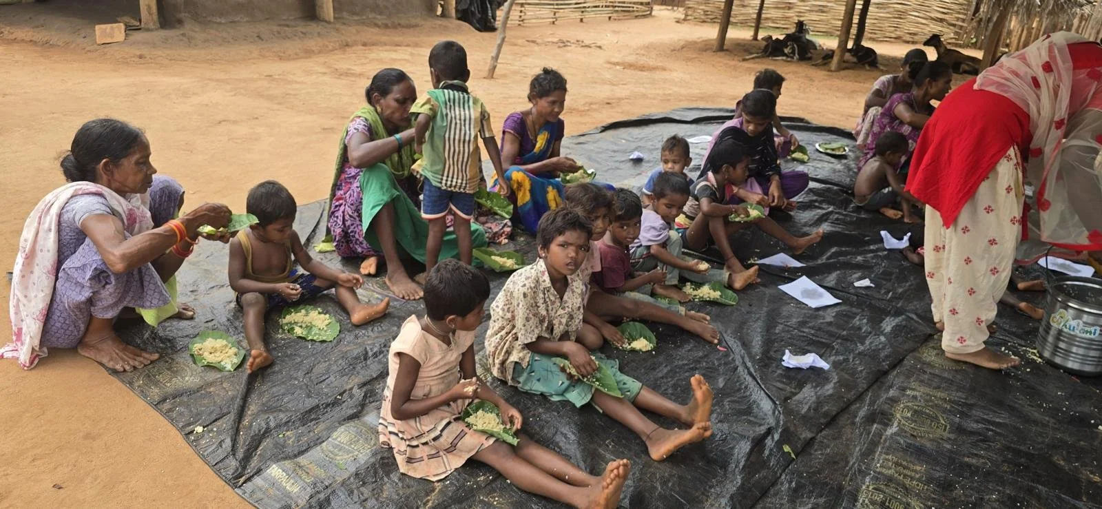 Group of women and children sitting on a black plastic sheet on the ground, eating food from banana leaves in an outdoor setting.
