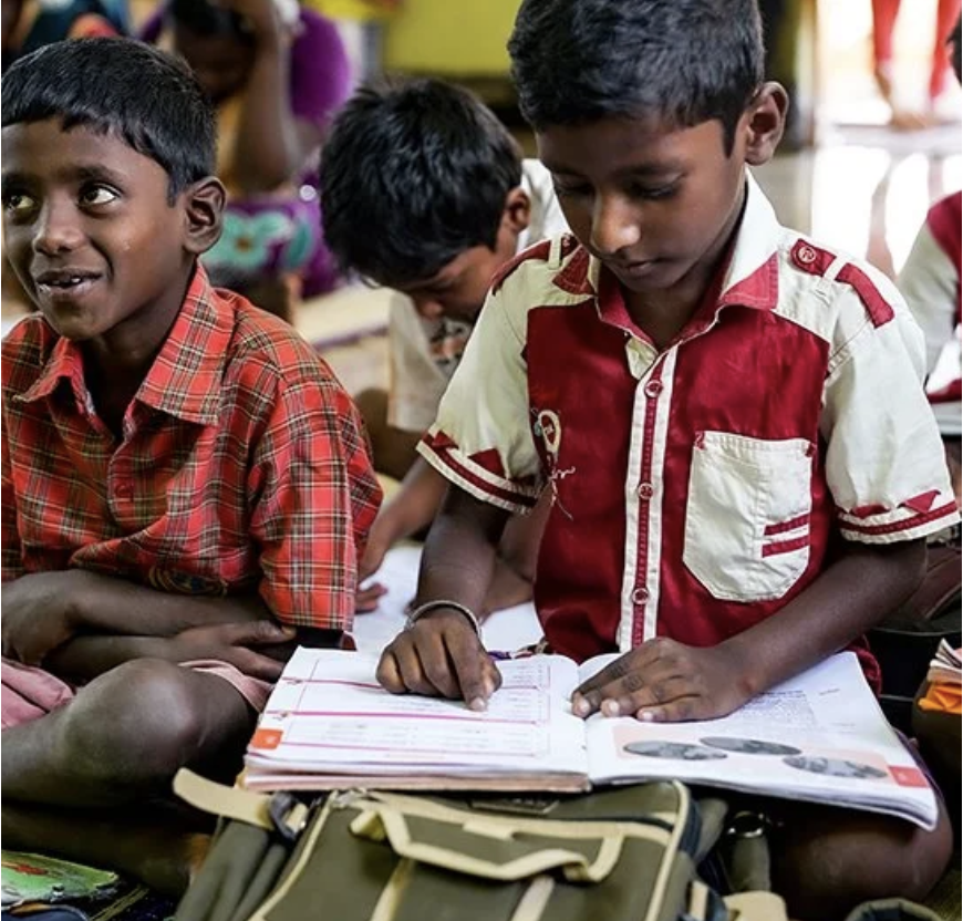 Two young boys sitting on the floor in a classroom, with one boy reading a book and the other boy smiling, wearing colorful shirts.