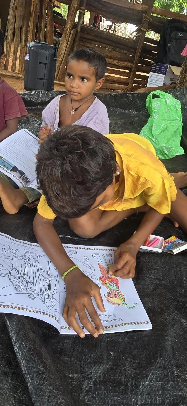 Two children are sitting at a table, one is coloring a picture of a dragon with crayons while the other watches. The setting appears to be outdoors with wooden structures in the background.