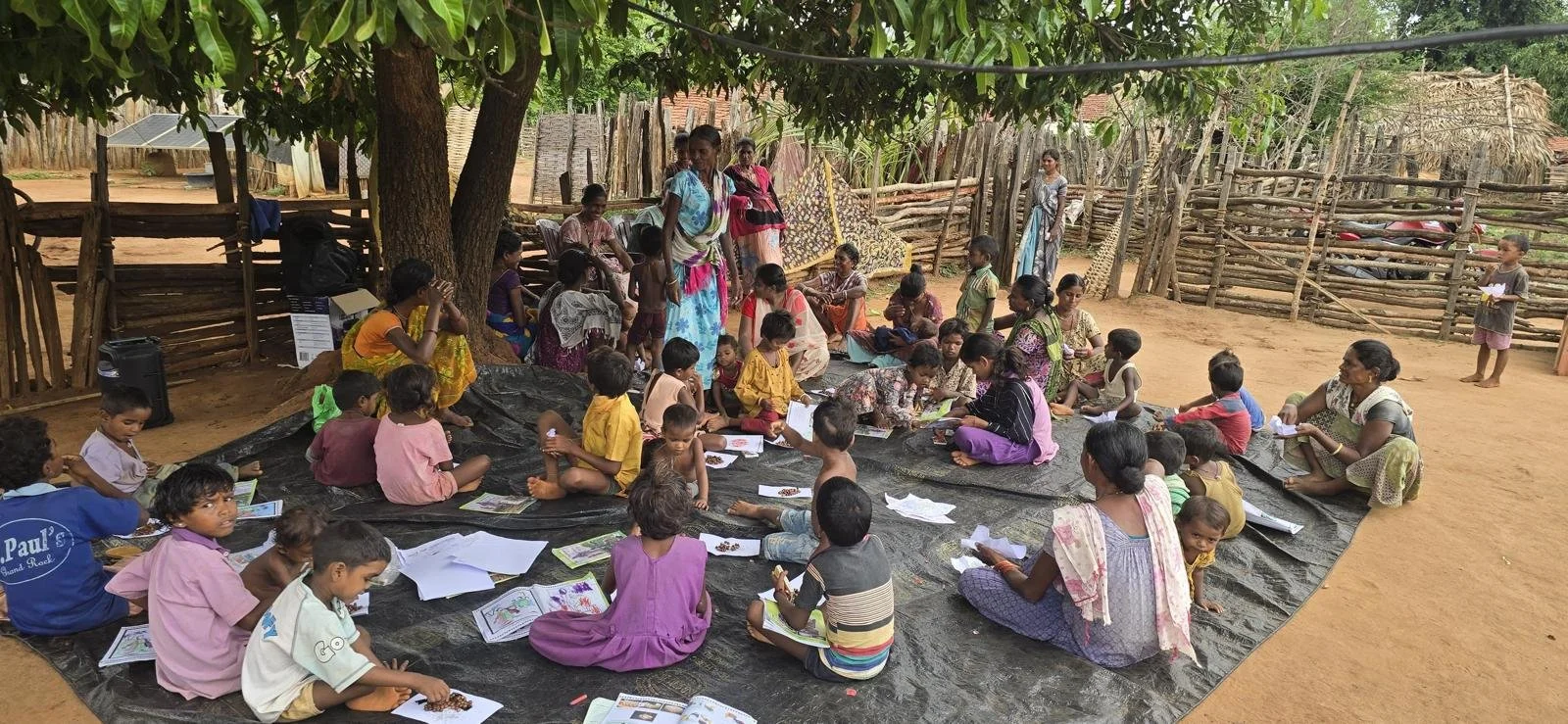 Children and women sitting on a black plastic sheet on the ground under a tree, engaging in educational activities, with a wooden fence and rustic buildings surrounding them.