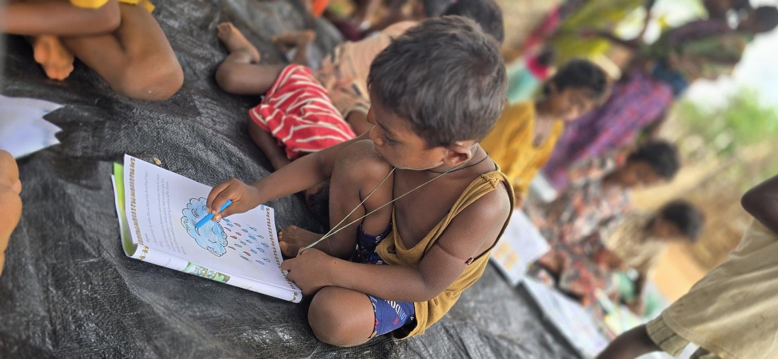 A young boy without a shirt, wearing shorts and a necklace, sitting on a black mat and coloring in a workbook with a blue pen, while other children and adults sit and stand nearby in the background.