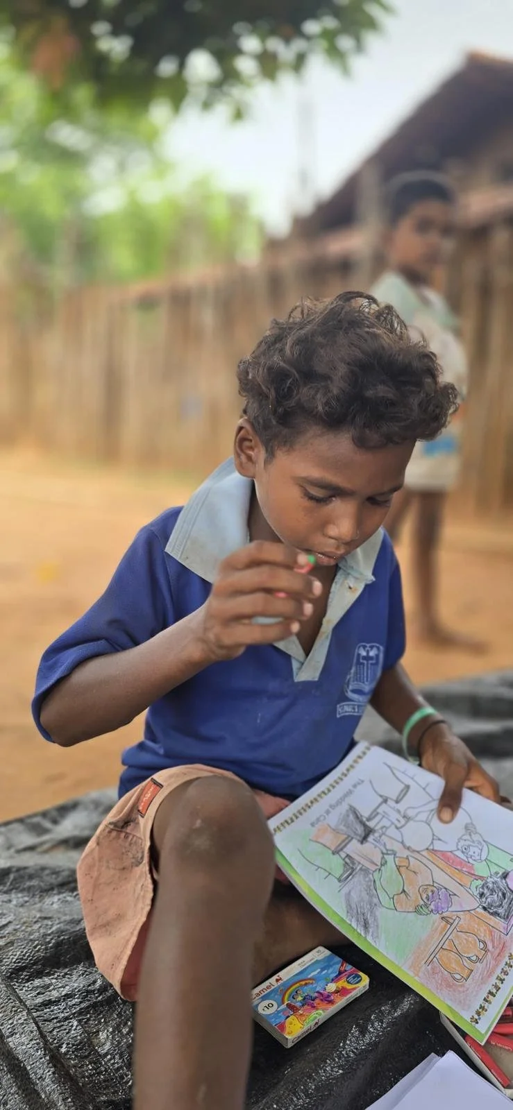 A young boy with curly hair wearing a blue school uniform sitting outdoors, coloring in a book while holding a crayon, with a second person blurred in the background.