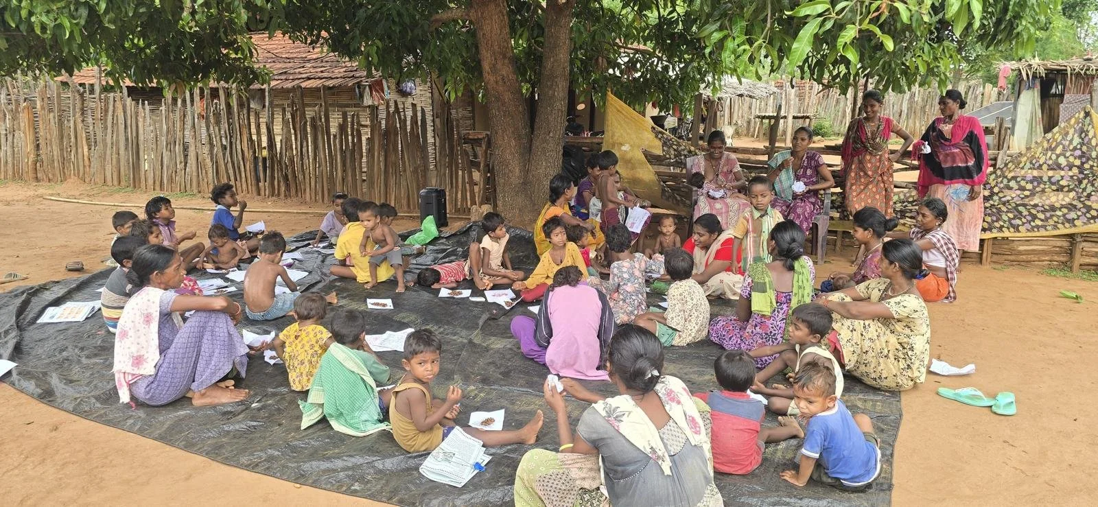 A large group of children and women gathered outdoors under a tree, seated on a black tarpaulin with papers and books, participating in an outdoor learning activity in a rural area.