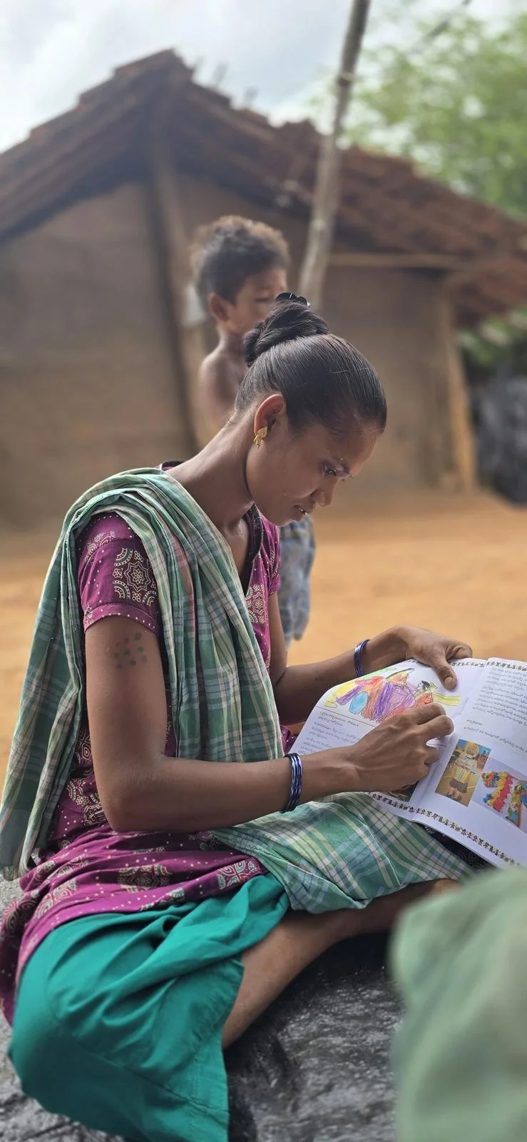 Woman sitting cross-legged on the ground reading a colorful illustrated book with a village house and a child standing behind her.