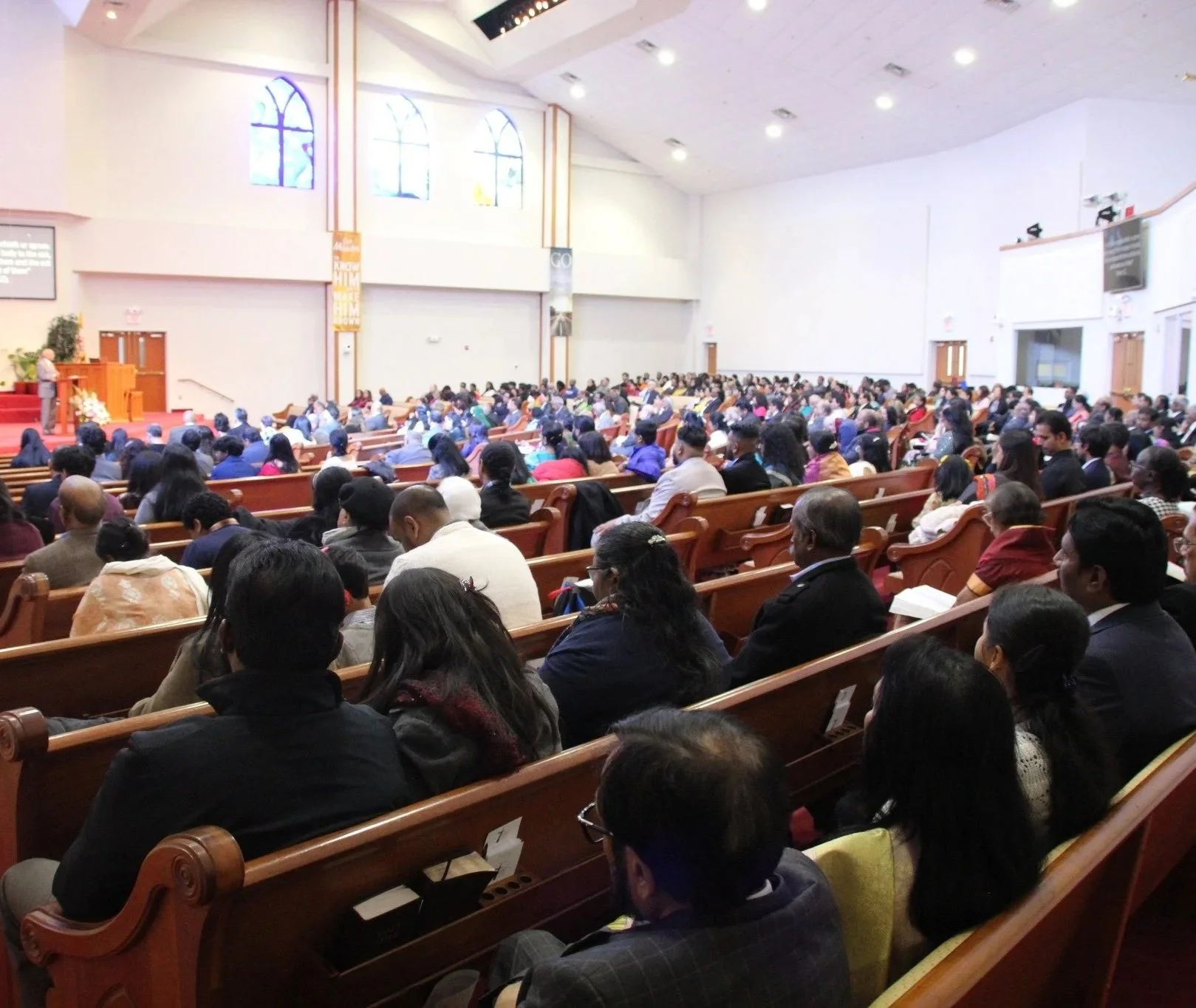 Inside a large church or auditorium filled with a diverse congregation of people seated in wooden pews, listening to a speaker at the podium.
