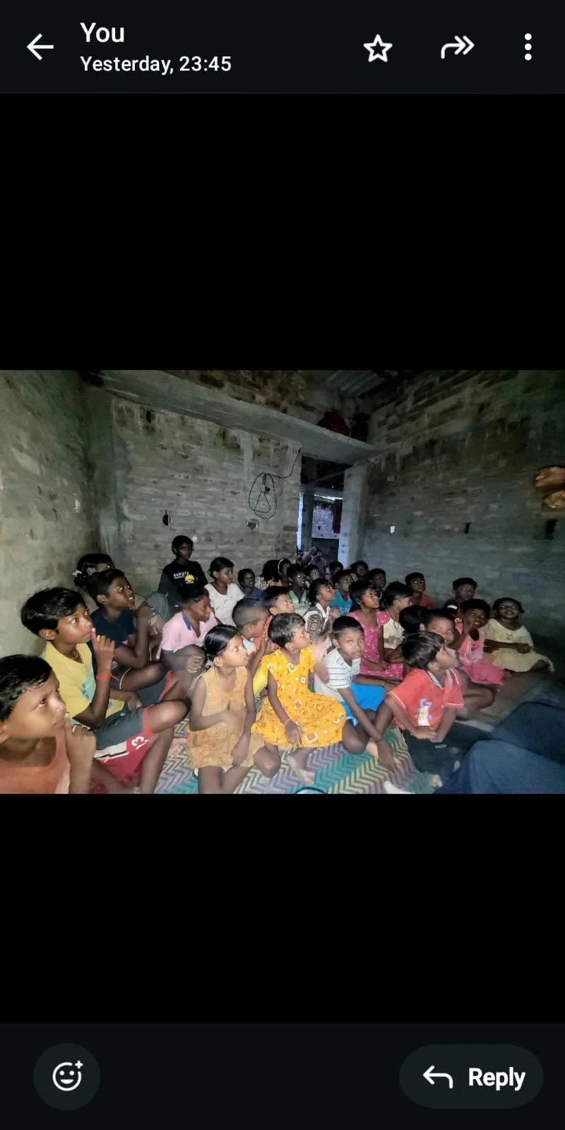 Group of children sitting on the floor inside a rustic, unfinished brick room, attentively watching something out of the frame.